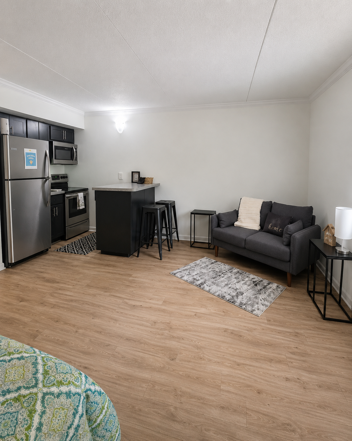 A modern, open-concept living space featuring a kitchen with stainless steel appliances, a grey sofa, a small dining area with bar stools, and light wood flooring. Soft lighting and decorative accents add warmth, while a patterned rug adds texture. The design is minimal and inviting.