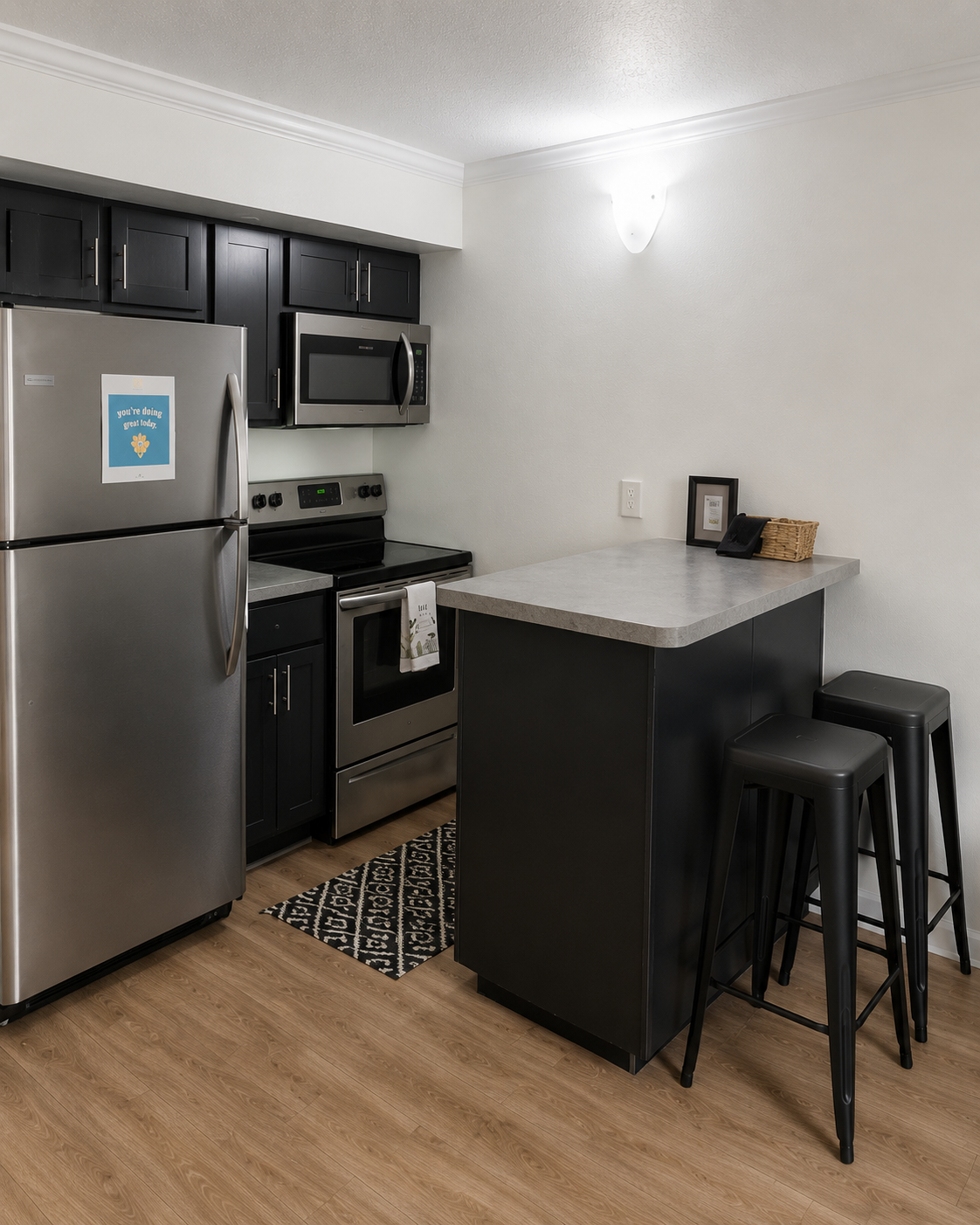 Modern kitchen featuring dark cabinetry, stainless steel appliances including a refrigerator and oven, and a concrete countertop. A small island with two black stools is prominent. The room has a light-colored wall and a decorative rug on the floor, creating a stylish and functional space.