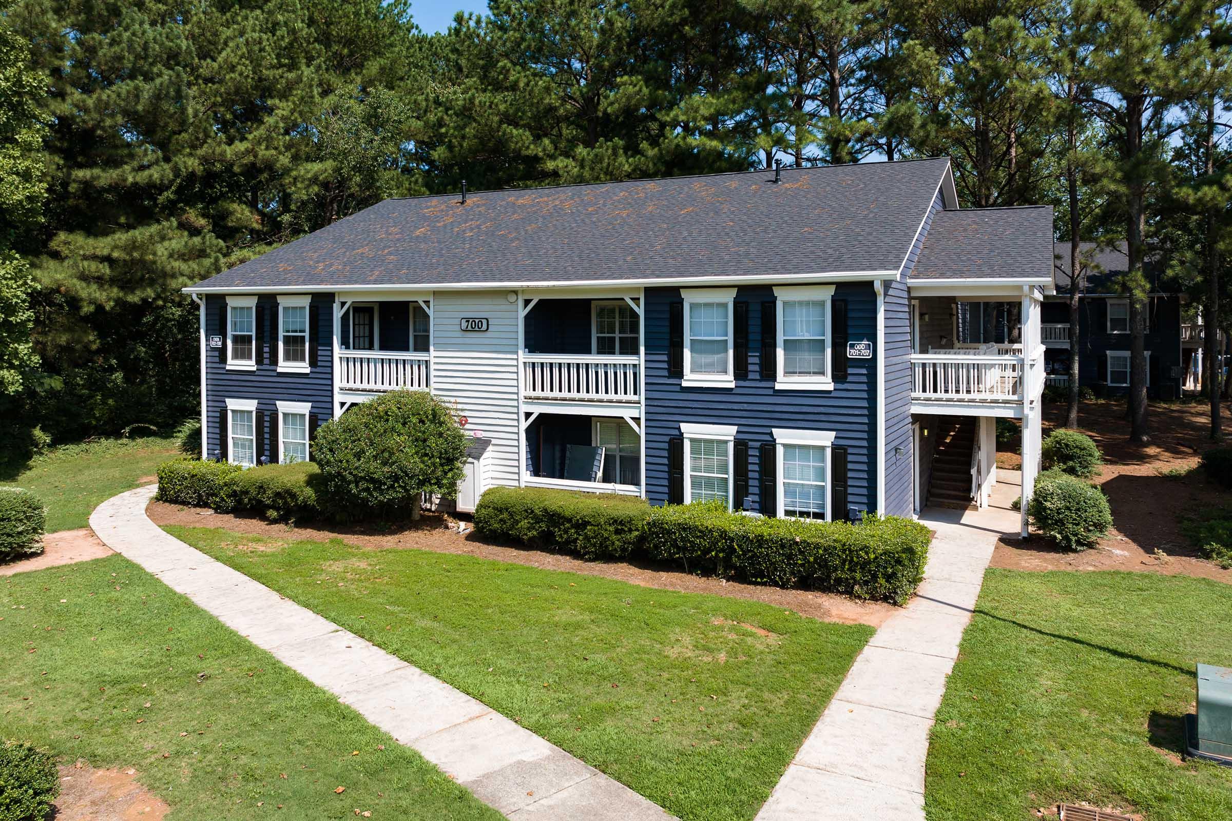 Two-story residential building with blue siding and white trim, surrounded by green grass and neatly trimmed bushes. A concrete walkway leads to the entrance, and tall trees are visible in the background under a clear blue sky.