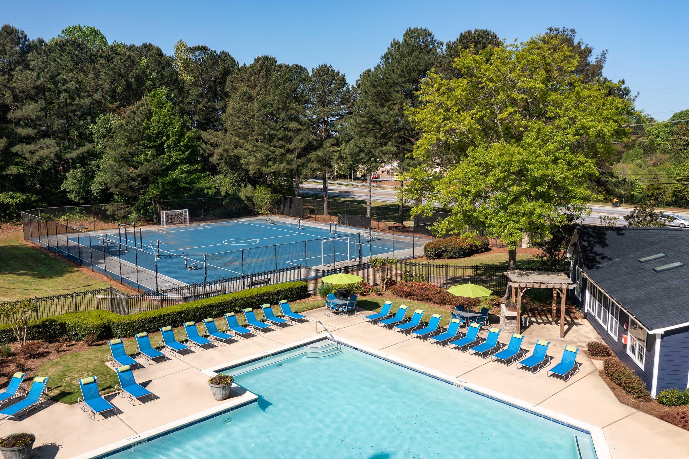 A bright outdoor area featuring a swimming pool with lounge chairs, surrounded by neatly landscaped greenery. In the background, a multi-use sports court is visible, situated next to tall trees under a clear blue sky.