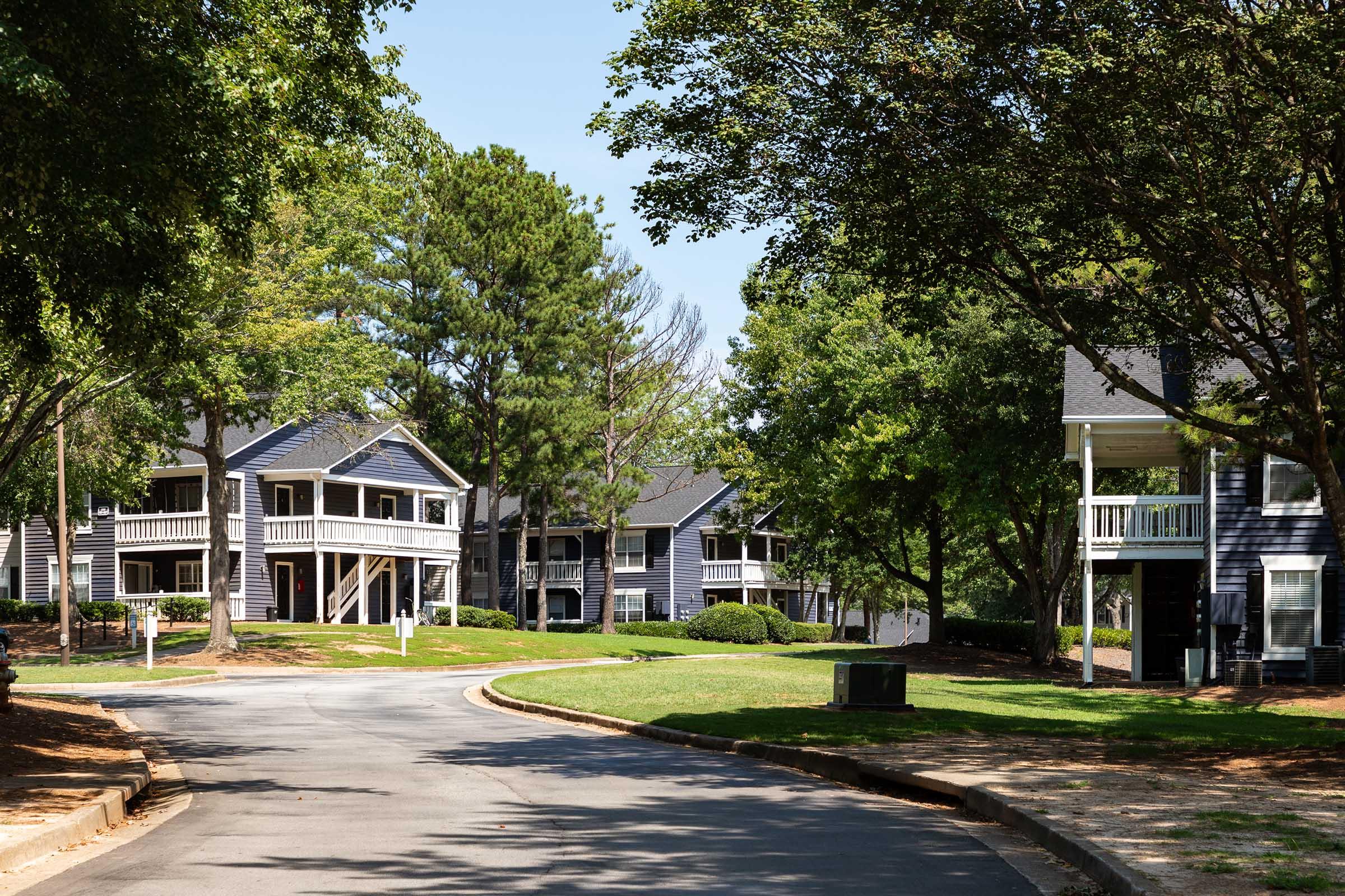 A quiet residential neighborhood featuring two-story houses with dark blue siding and white trim. The street curves through the area, lined with green lawns and trees, creating a peaceful atmosphere under a clear blue sky.