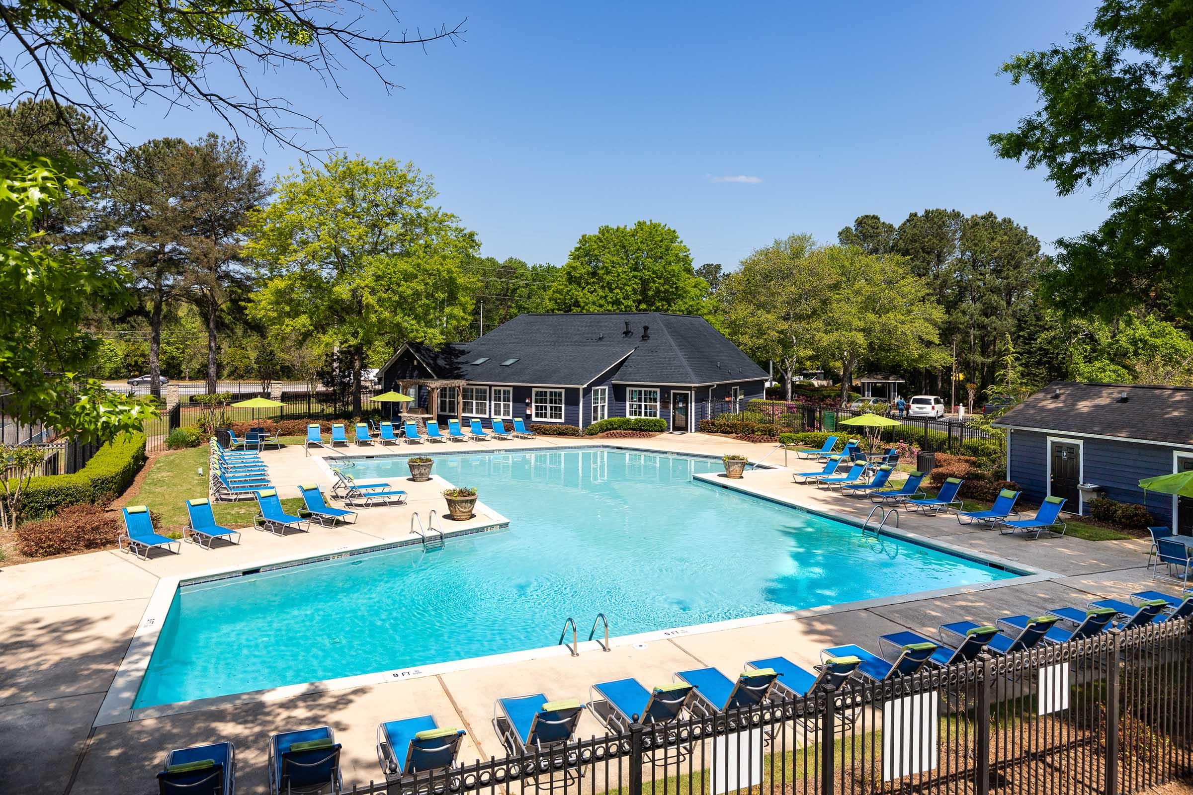 A large outdoor swimming pool surrounded by green trees and lounge chairs. There's a clubhouse in the background, and a small shed to the side. The scene is bright and sunny, with clear blue skies.