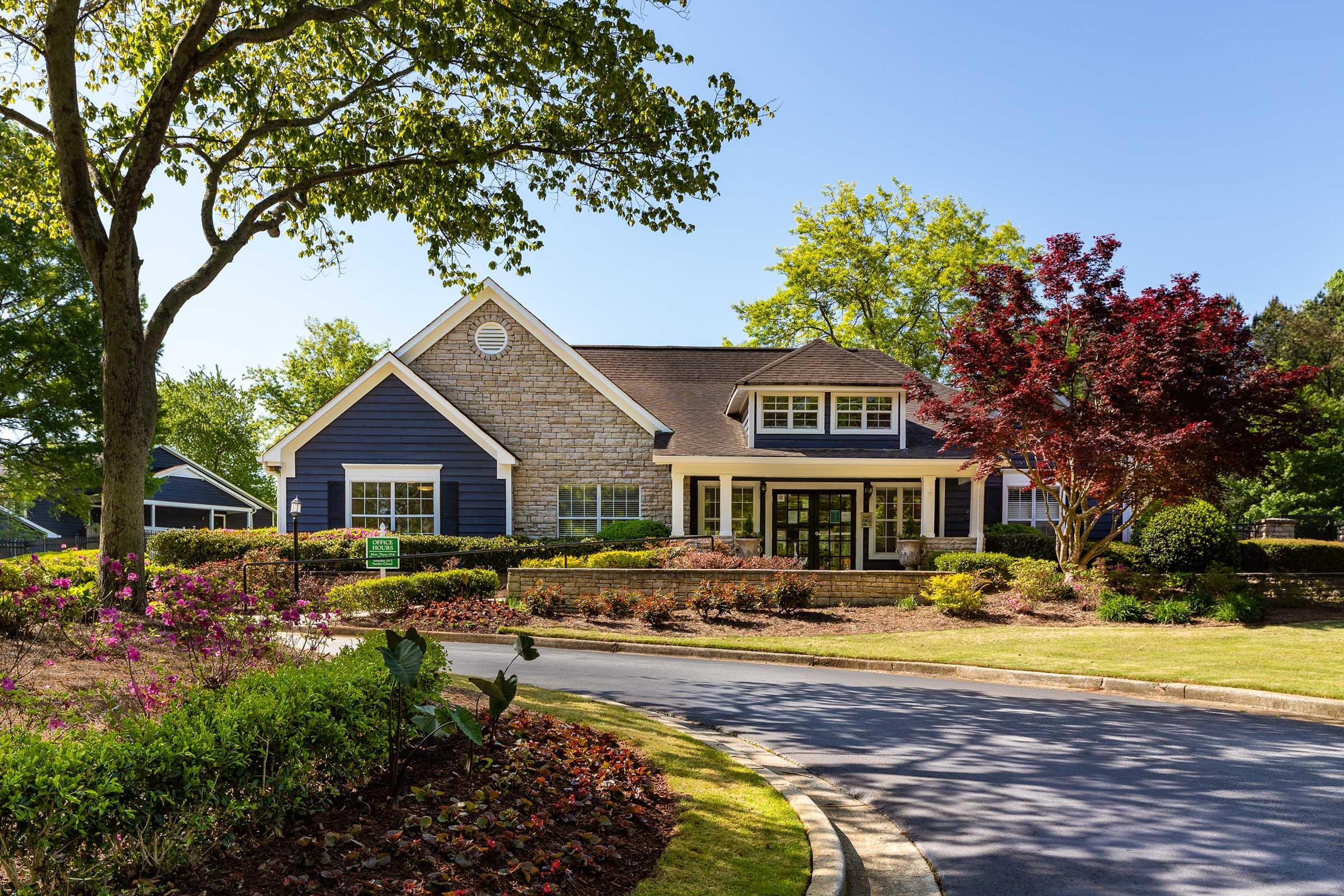 A beautiful suburban house featuring a mix of stone and blue siding, surrounded by well-maintained landscaping. The front yard includes shrubs, flowering plants, and trees, with a clear blue sky overhead. The driveway curves gently, leading to the entrance of the home.