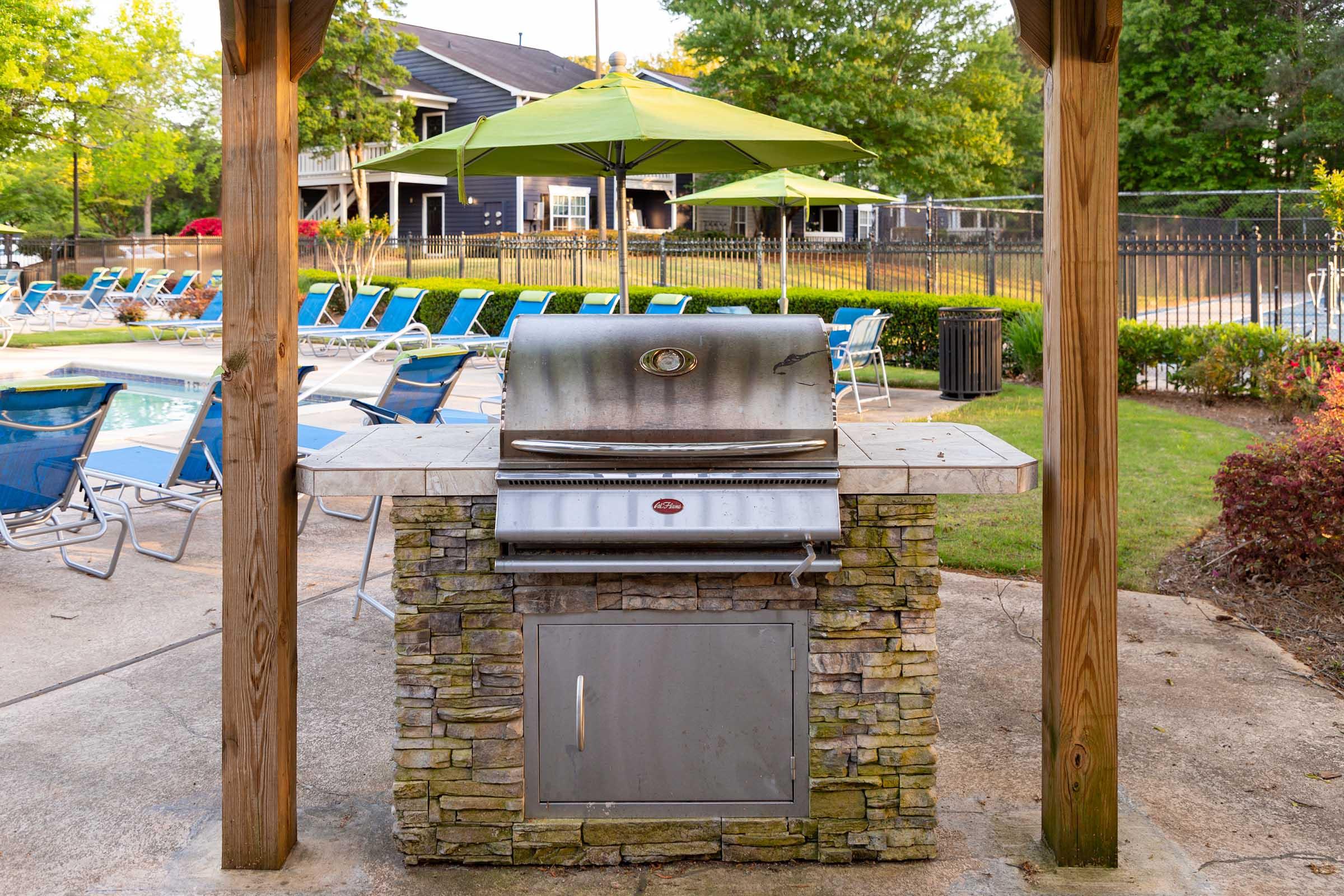 An outdoor grilling area featuring a stainless steel grill on a stone platform. There are blue lounge chairs arranged around a swimming pool in the background, with green umbrellas providing shade. Lush landscaping and a fenced area can be seen, creating a relaxing atmosphere.