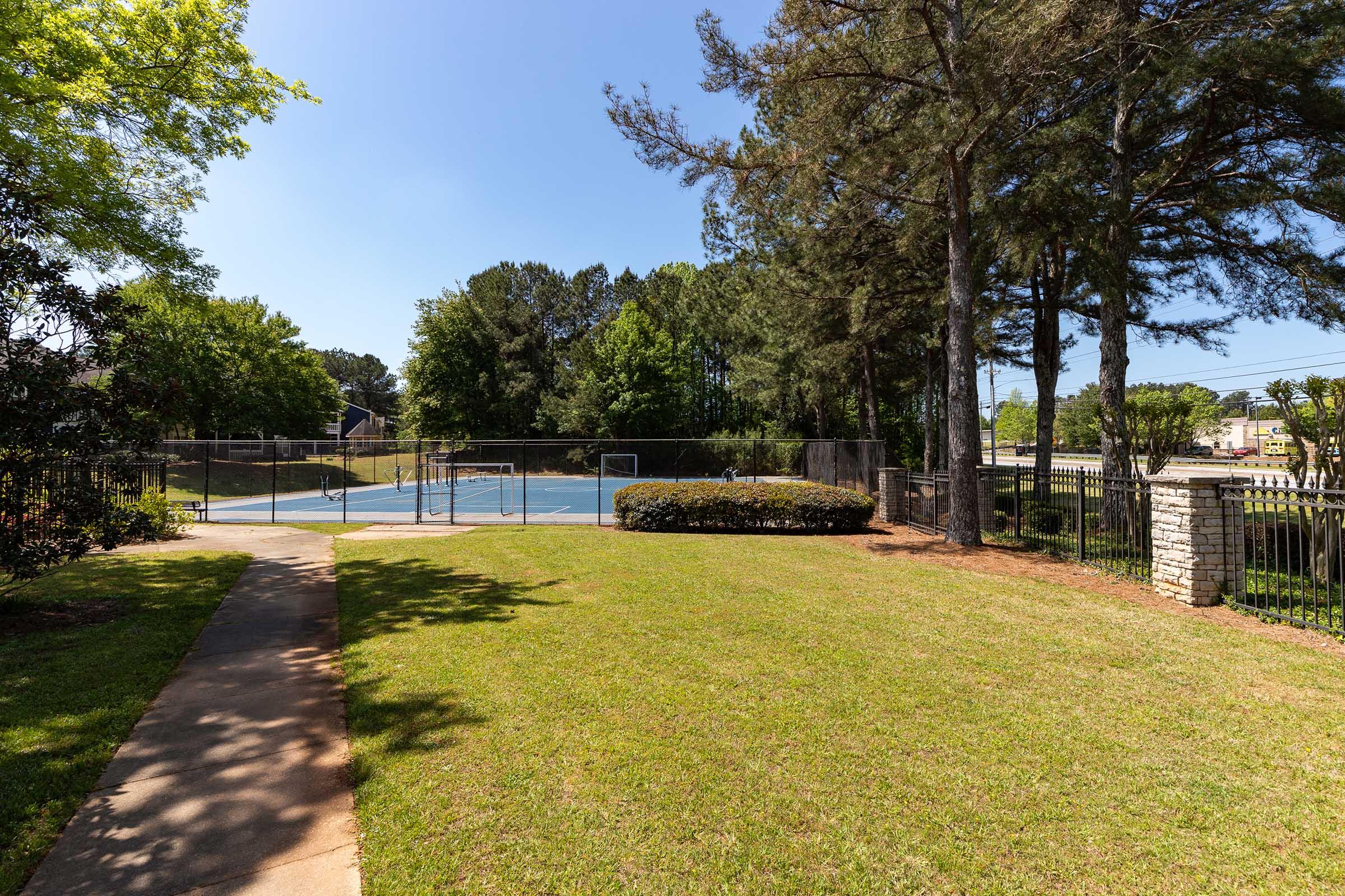 A well-maintained outdoor area featuring a grassy lawn, a walkway, and a fenced tennis court in the background. Tall trees provide shade, while a few shrubs are neatly trimmed along the path. The scene is bright and sunny, indicating a pleasant day.