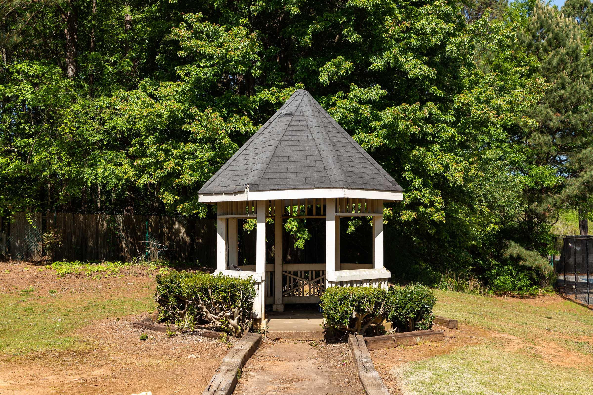 A white gazebo with a black shingle roof sits in a grassy area surrounded by lush green trees. The gazebo features an open design with benches inside and a pathway leading to it, inviting relaxation in a tranquil outdoor setting.