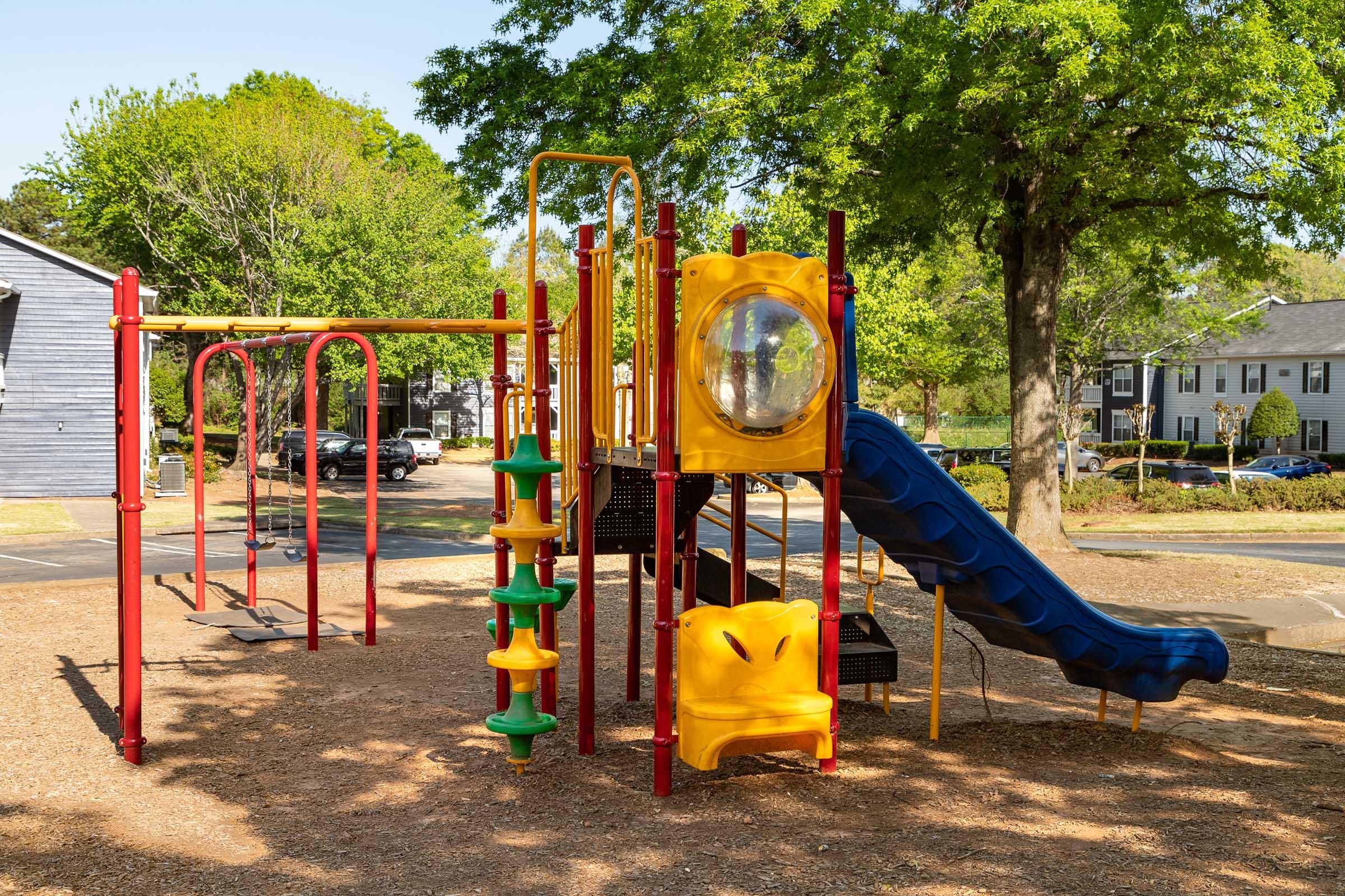 Colorful playground equipment featuring a yellow slide, swings, and various climbing structures set in a grassy area surrounded by trees and residential buildings. The scene captures a sunny day, emphasizing the inviting atmosphere for children to play.