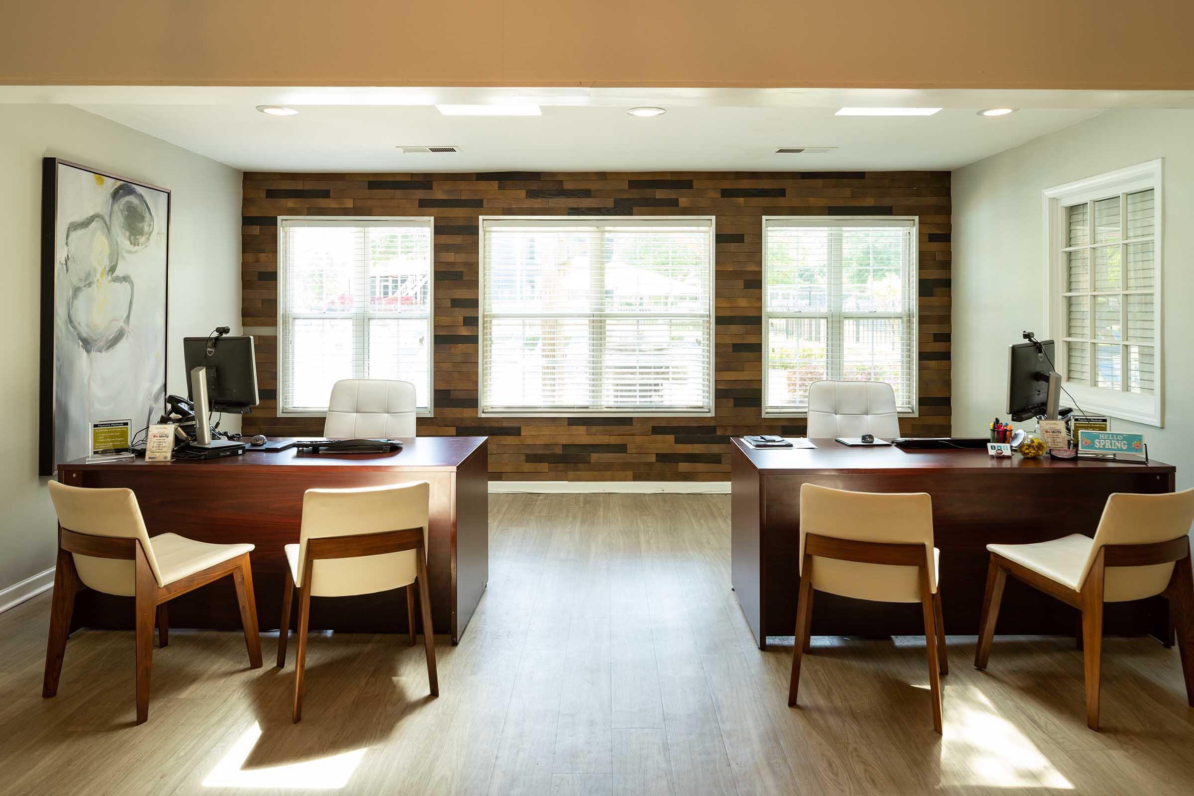Two modern office desks facing each other in a bright, inviting office space. Each desk is equipped with a computer and office supplies. Large windows allow natural light to fill the room, highlighting wooden accent walls. Four light-colored chairs are positioned around the desks, creating a professional atmosphere.