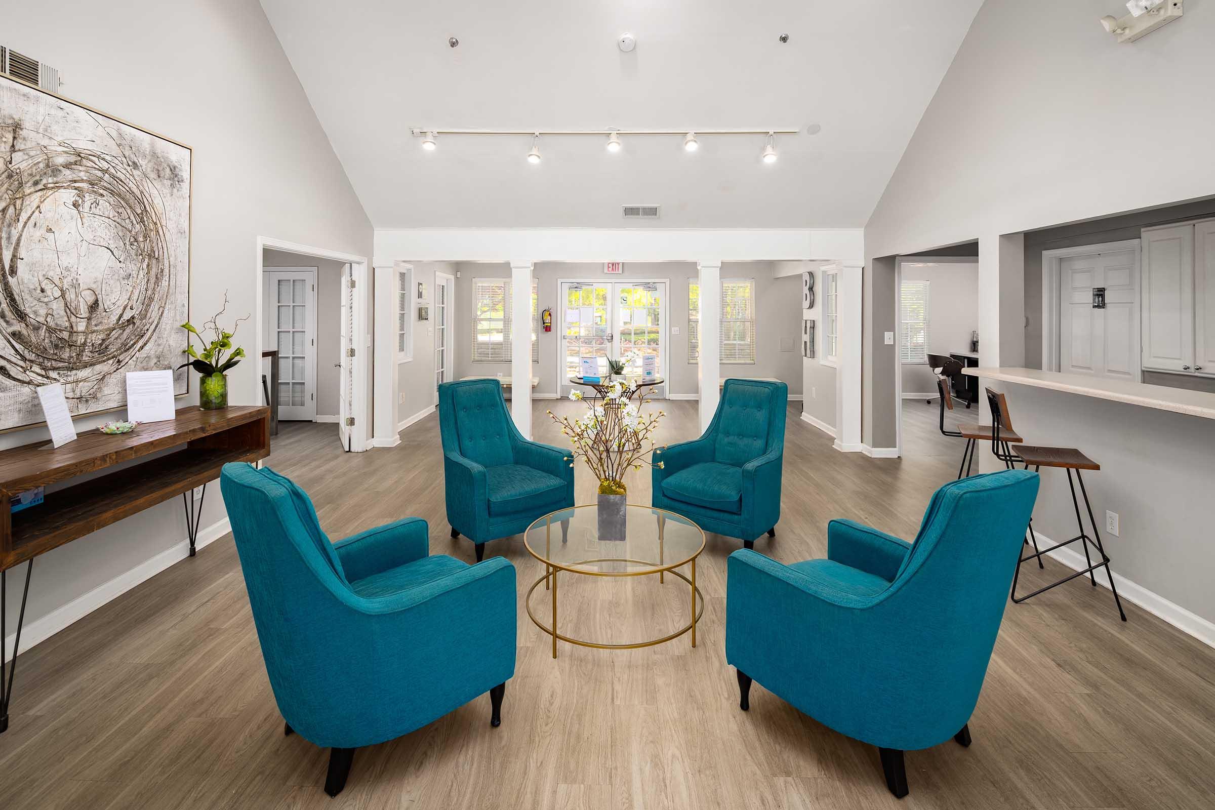 A modern lobby featuring four turquoise chairs arranged around a glass coffee table. The walls are light-colored, and there are large windows allowing natural light. A wooden console table holds decorative items on one side, and there's a bar-style seating area on the other.