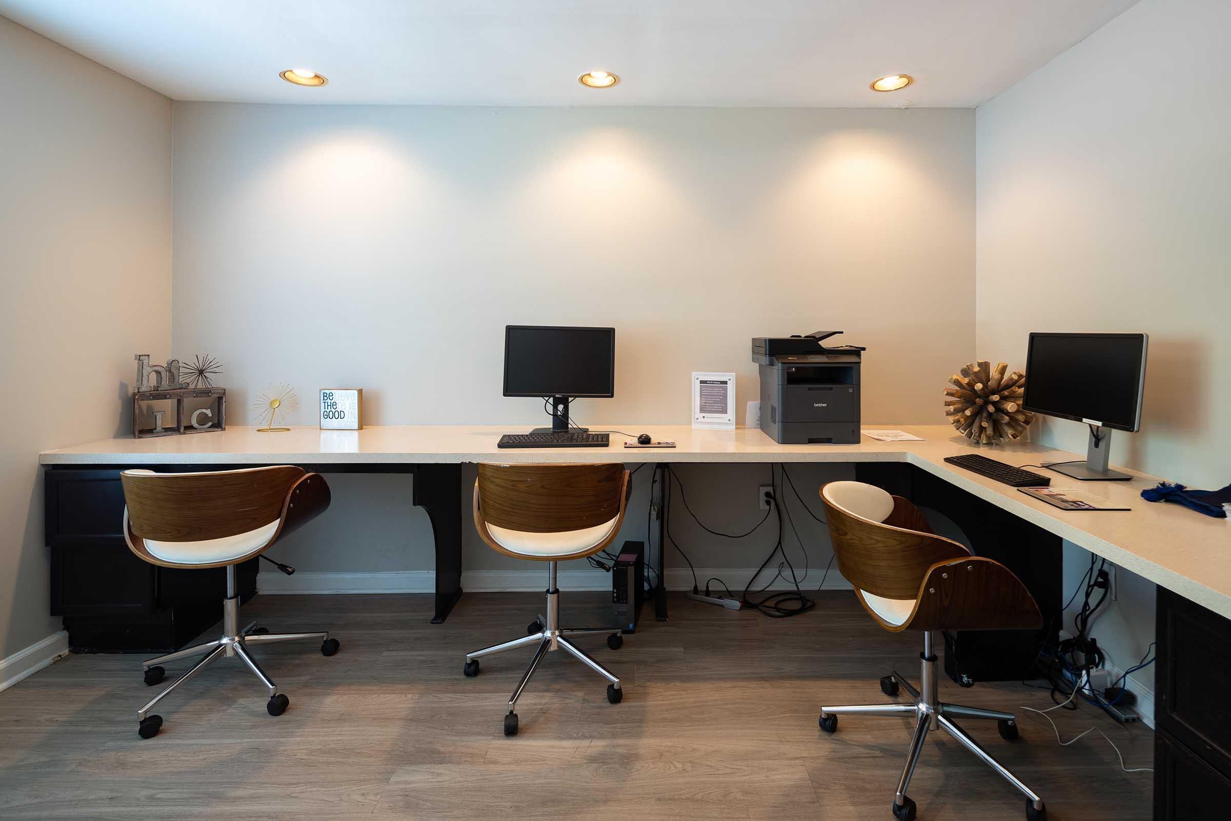 A modern workspace with two computer workstations, each featuring a monitor and a keyboard. There are three stylish wooden office chairs with white seats. A printer is located on the left side of the desk. The wall is painted in a light color, and there are soft overhead lights illuminating the space.