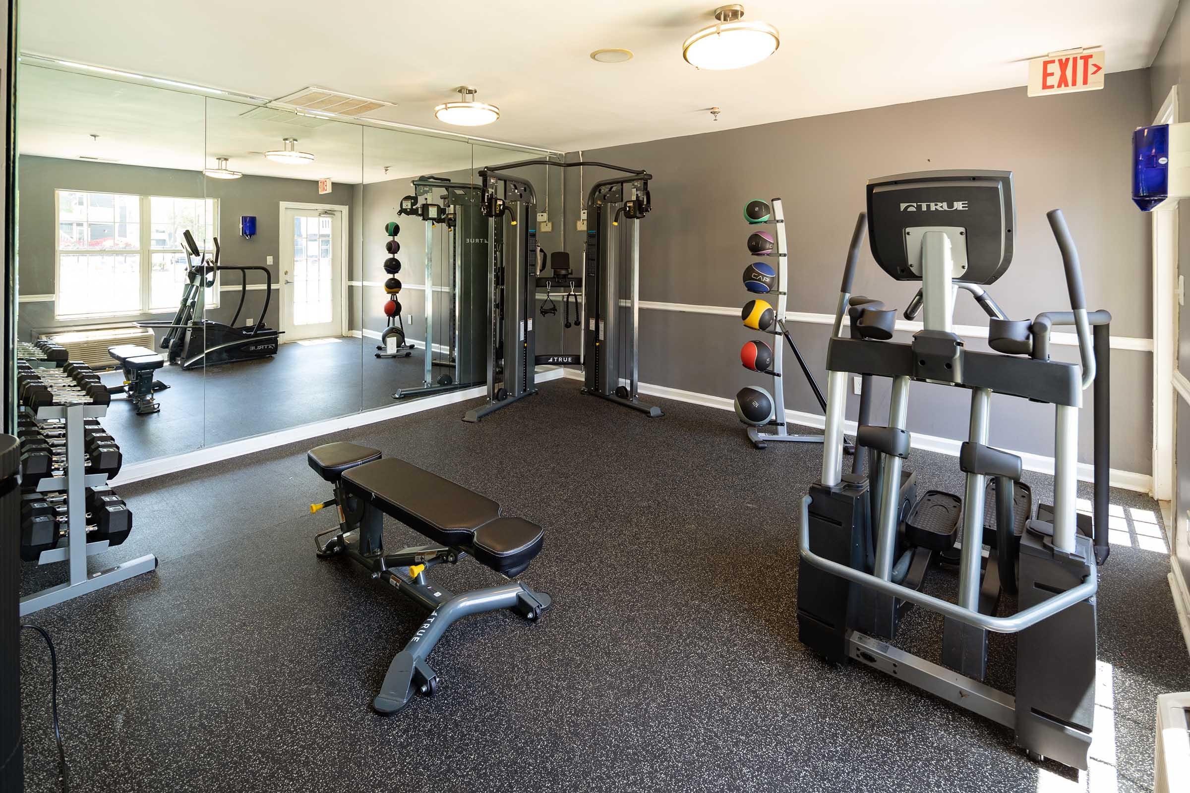 A modern gym interior featuring various workout equipment, including weight benches, dumbbells, and cable machines. The floor is black rubber, and a large mirror reflects the space. Bright lighting enhances the atmosphere, with exercise balls neatly arranged on a rack.