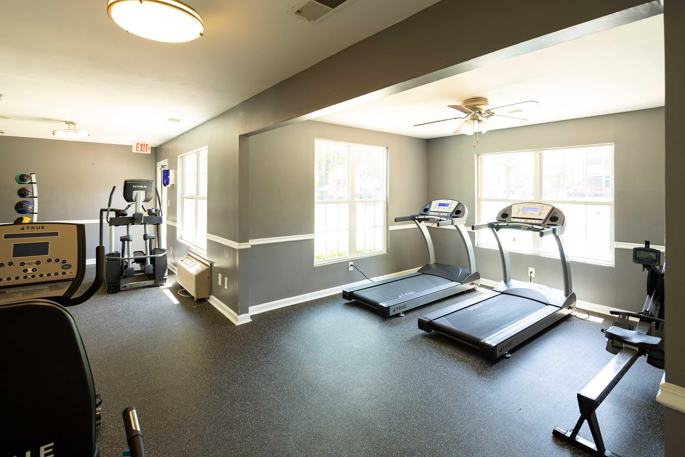 A bright and spacious gym area with two treadmills facing large windows, a ceiling fan, and fitness equipment including an elliptical machine and weight station. The flooring is dark and there is ample natural light from the windows, creating an inviting atmosphere for exercise.