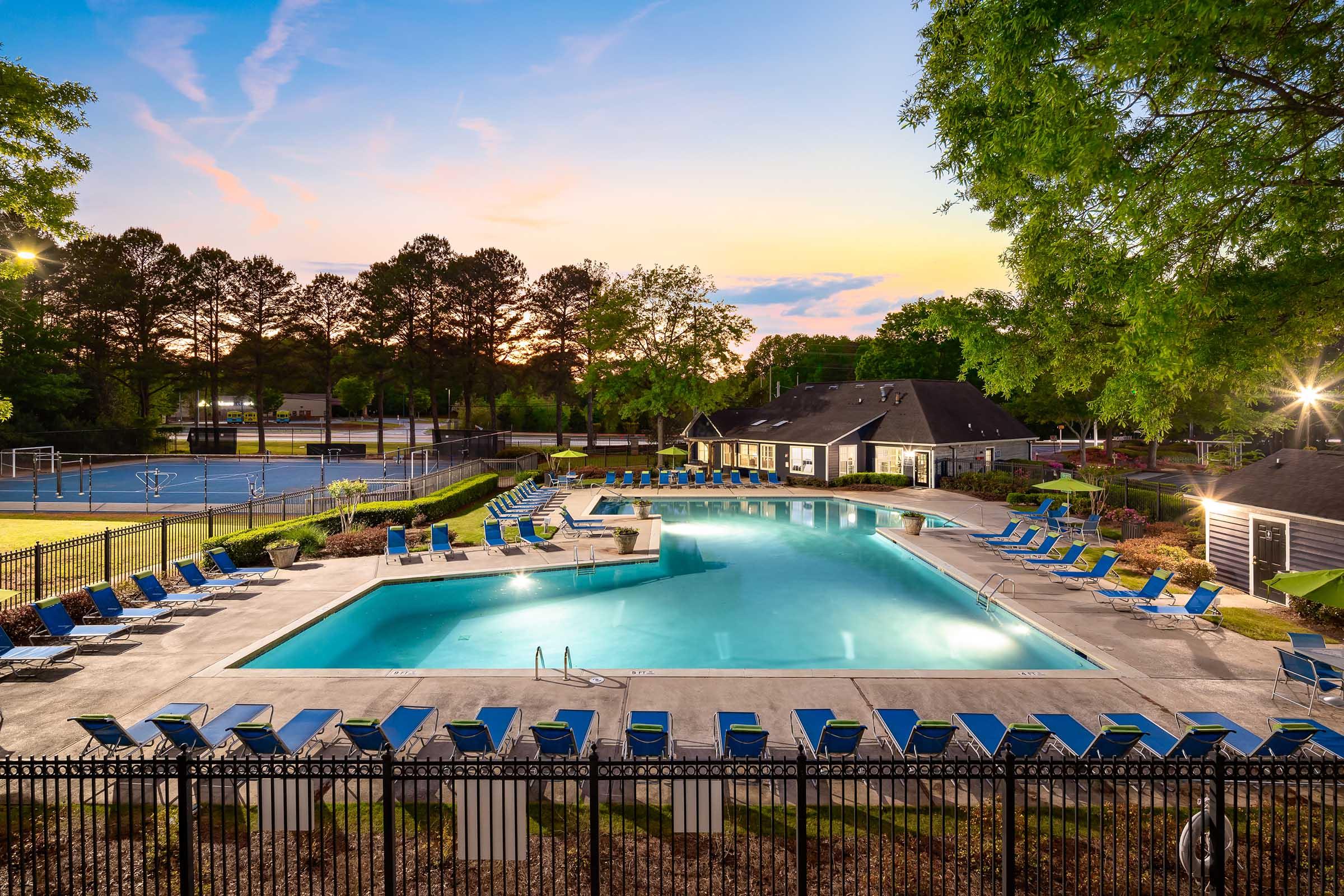 A spacious outdoor swimming pool surrounded by lounge chairs, with a jacuzzi nearby. The area is beautifully landscaped with trees and grass, and there's a tennis court in the background. The scene is set at sunset, with a warm glow illuminating the pool area and surrounding amenities.