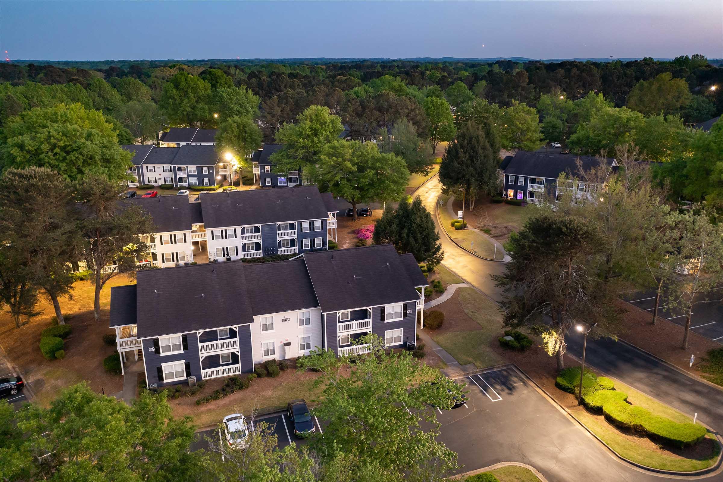 Aerial view of a residential complex at twilight, featuring several multi-story apartment buildings surrounded by green trees and well-maintained landscaping. Pathways and parking areas are visible, with soft lighting illuminating the scene, creating a serene atmosphere.