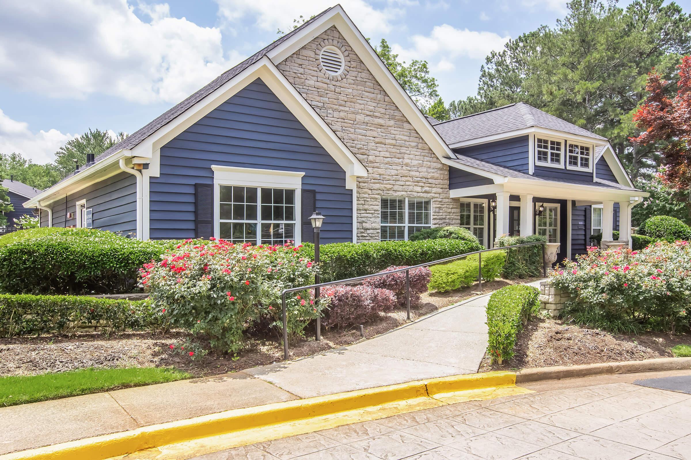 A beautiful blue and stone house surrounded by well-maintained gardens, featuring colorful flower beds and green shrubs. The path leading to the front door is bordered by plants, with tall trees providing a lush backdrop against a partly cloudy sky.