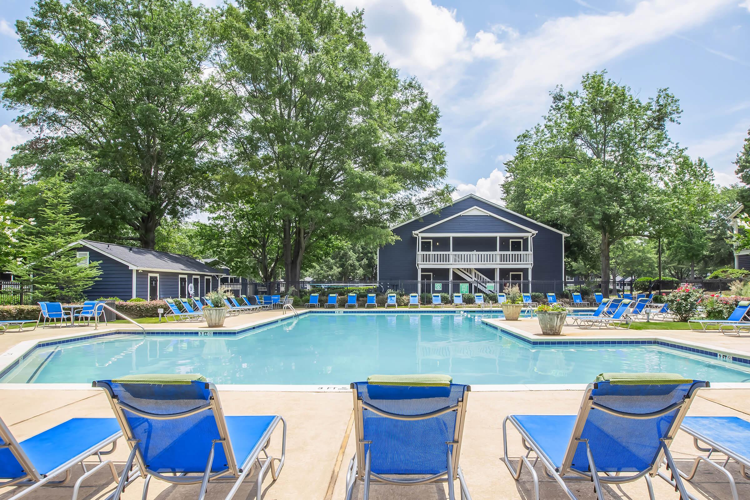A clear blue swimming pool surrounded by lounge chairs, with lush green trees in the background. A two-story building with a balcony overlooks the pool area, and the sky is bright with fluffy white clouds. The scene conveys a relaxed and inviting outdoor atmosphere.