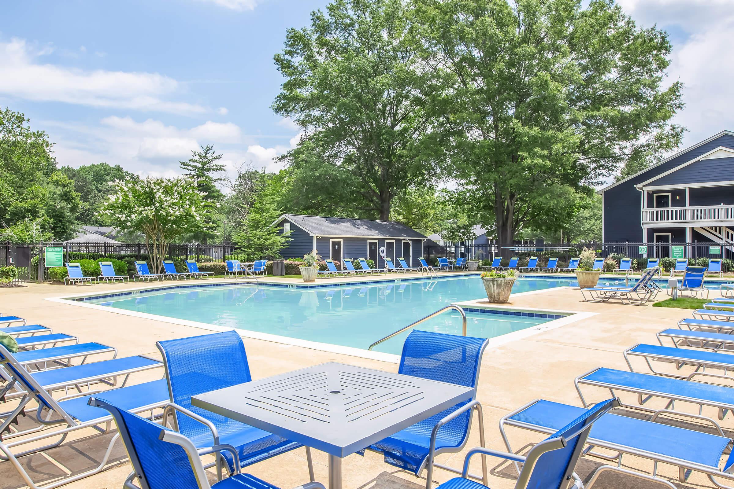 A sunny outdoor pool area surrounded by blue lounge chairs and tables. Lush green trees and a few buildings are in the background, creating a relaxing atmosphere. The pool is clear and inviting, ideal for leisure and recreation.