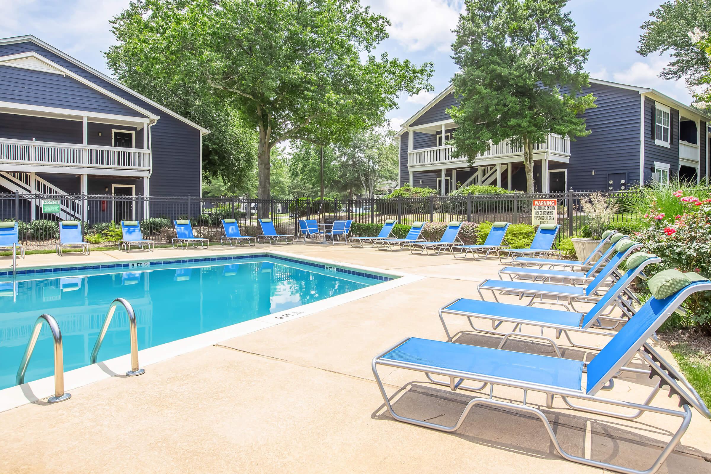 A clear swimming pool surrounded by blue lounge chairs and green trees. Two multi-story dark-colored apartment buildings are visible in the background, with a well-maintained landscape. The scene is bright and inviting, perfect for relaxation and enjoying outdoor leisure activities.
