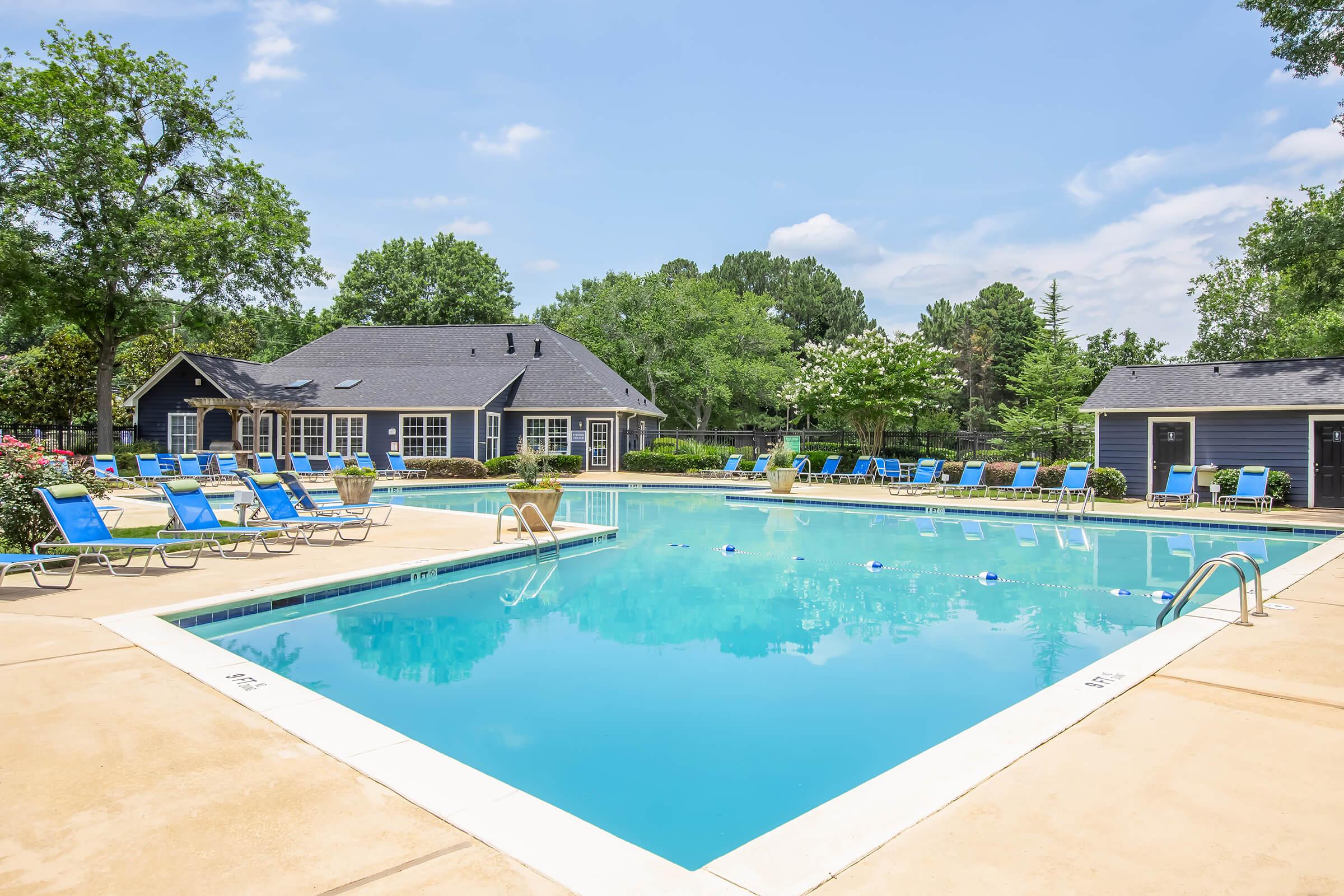 A clean, inviting outdoor swimming pool surrounded by lounge chairs under a clear blue sky. Lush greenery and trees provide a pleasant backdrop, with a clubhouse visible in the background, creating a relaxing atmosphere for leisure and enjoyment.