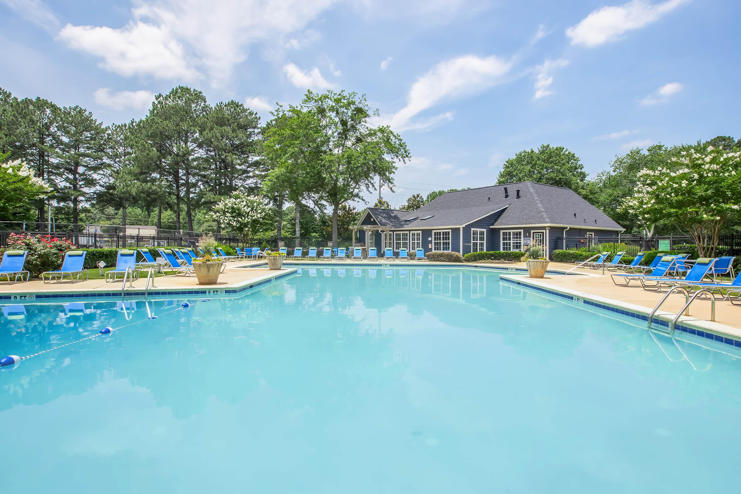 A large outdoor swimming pool surrounded by lounge chairs and potted plants. Lush green trees line the background, and a blue sky with scattered clouds completes the serene atmosphere. A clubhouse is visible nearby, creating a relaxing recreational space.