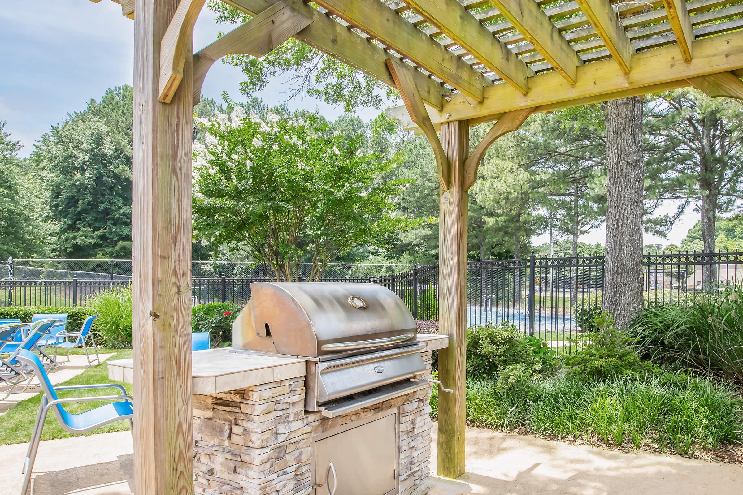 An outdoor grilling area with a stainless steel barbecue grill under a wooden pergola. Surrounding the grill are lounge chairs and lush greenery, with a swimming pool visible in the background, framed by trees and a fence. The scene suggests a relaxing outdoor space for cooking and leisure.
