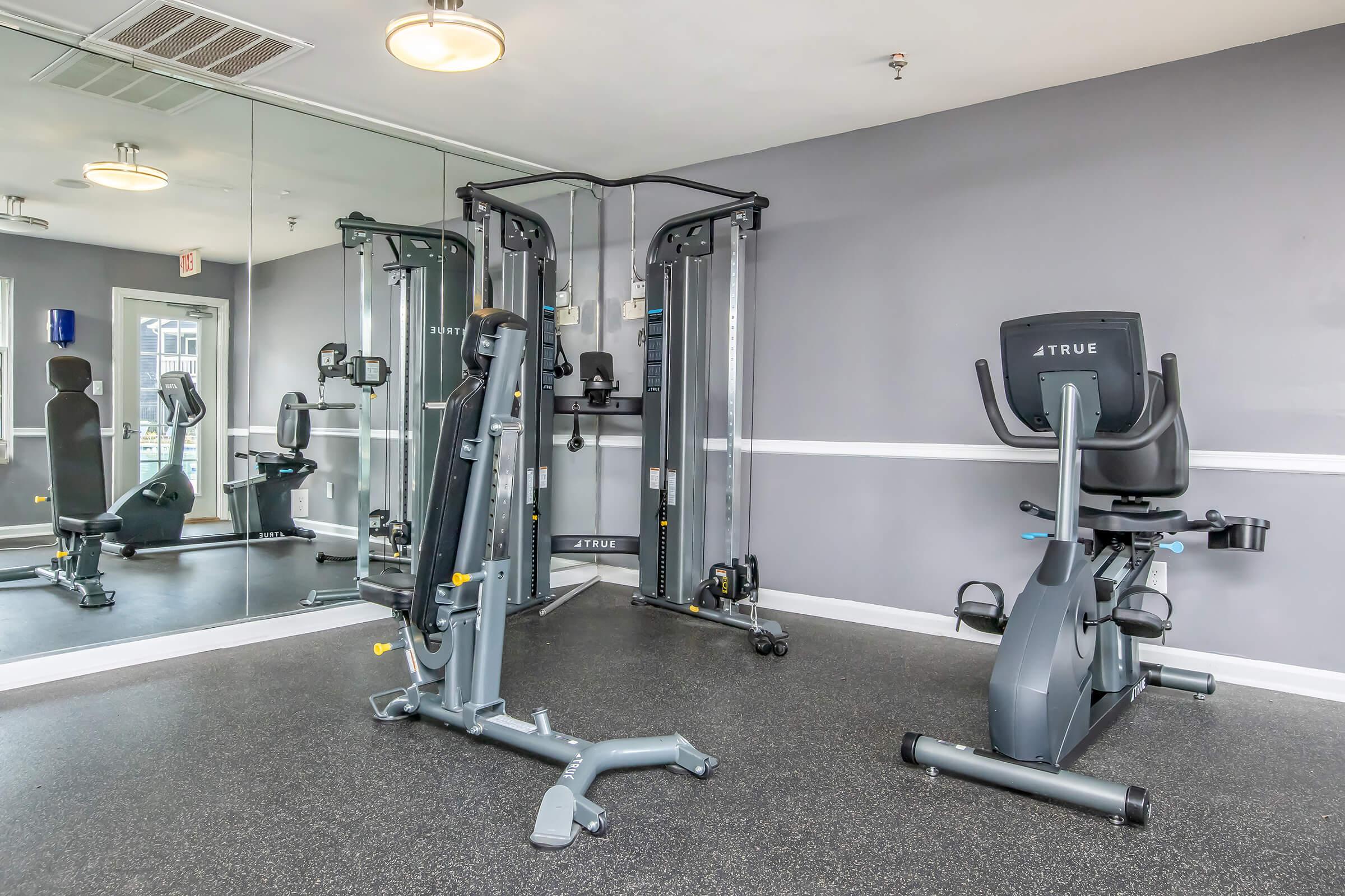 Interior of a modern gym featuring exercise equipment, including a stationary bike and a multi-station weight machine. The wall is painted gray, and there's a large mirror reflecting the equipment. The space is well-lit and designed for fitness activities.