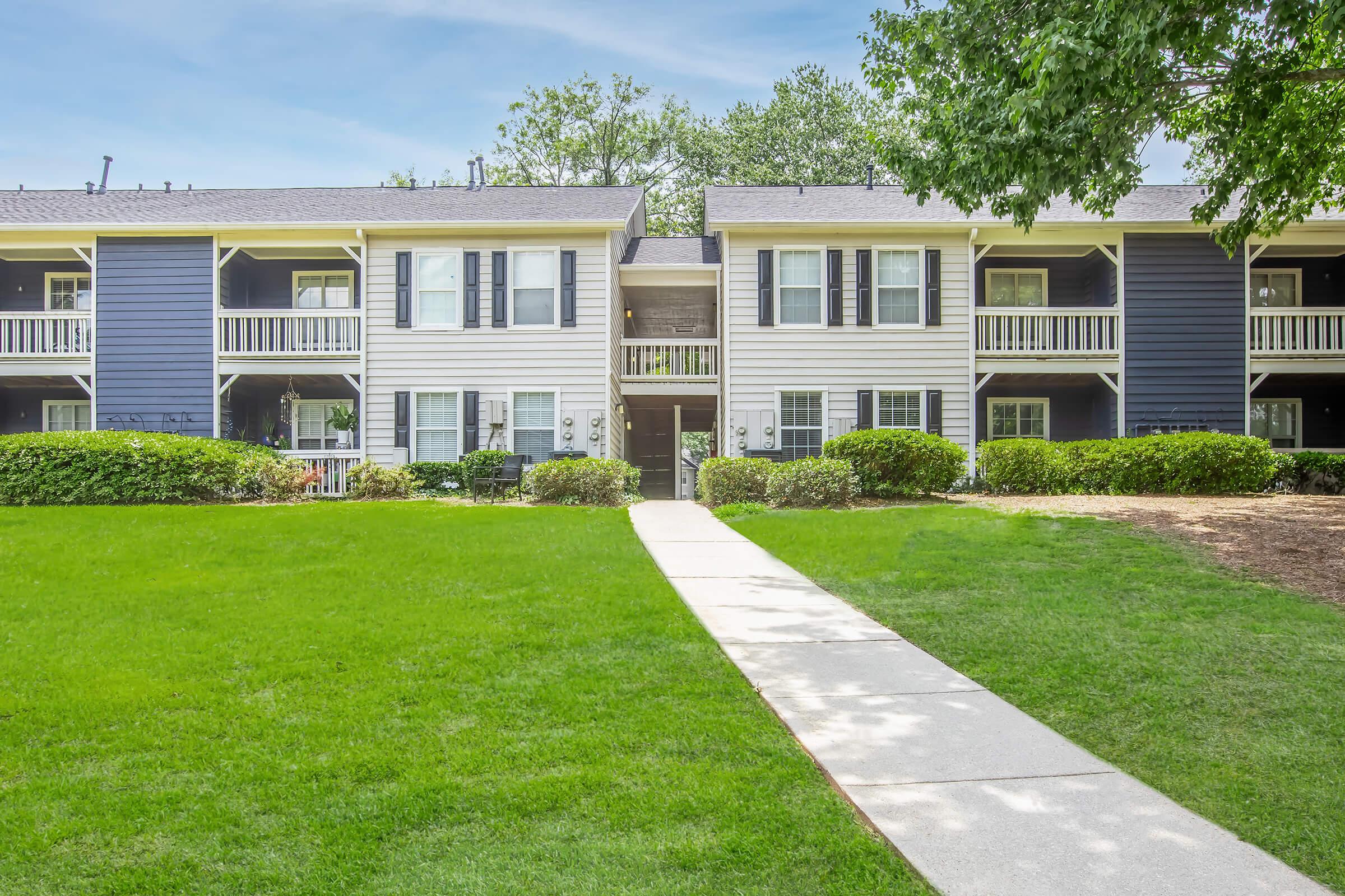 A two-story apartment building with a mix of blue and white siding. It has balconies on the upper level and a landscaped front yard with green grass and shrubs. A concrete walkway leads to the entrance, framed by well-maintained plants. The sky is clear and bright, adding to the inviting atmosphere.