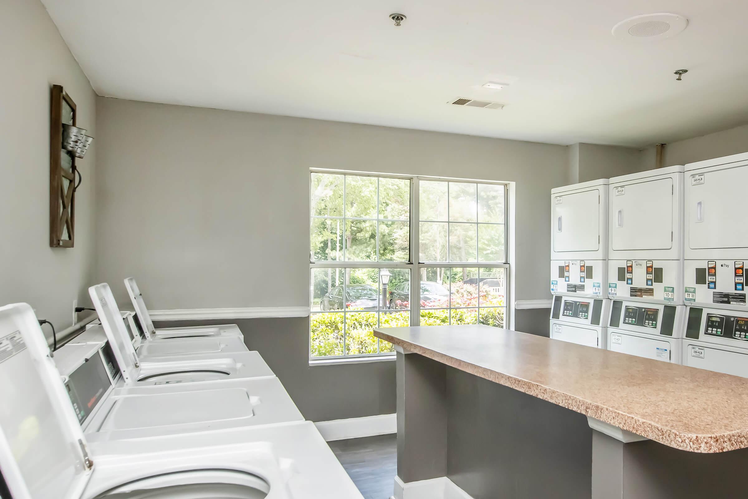A laundry room featuring several white washing machines and dryers lined up against a gray wall. A countertop is positioned in the center, with a large window allowing natural light to enter, revealing greenery outside. The atmosphere is clean and well-lit, ideal for doing laundry.