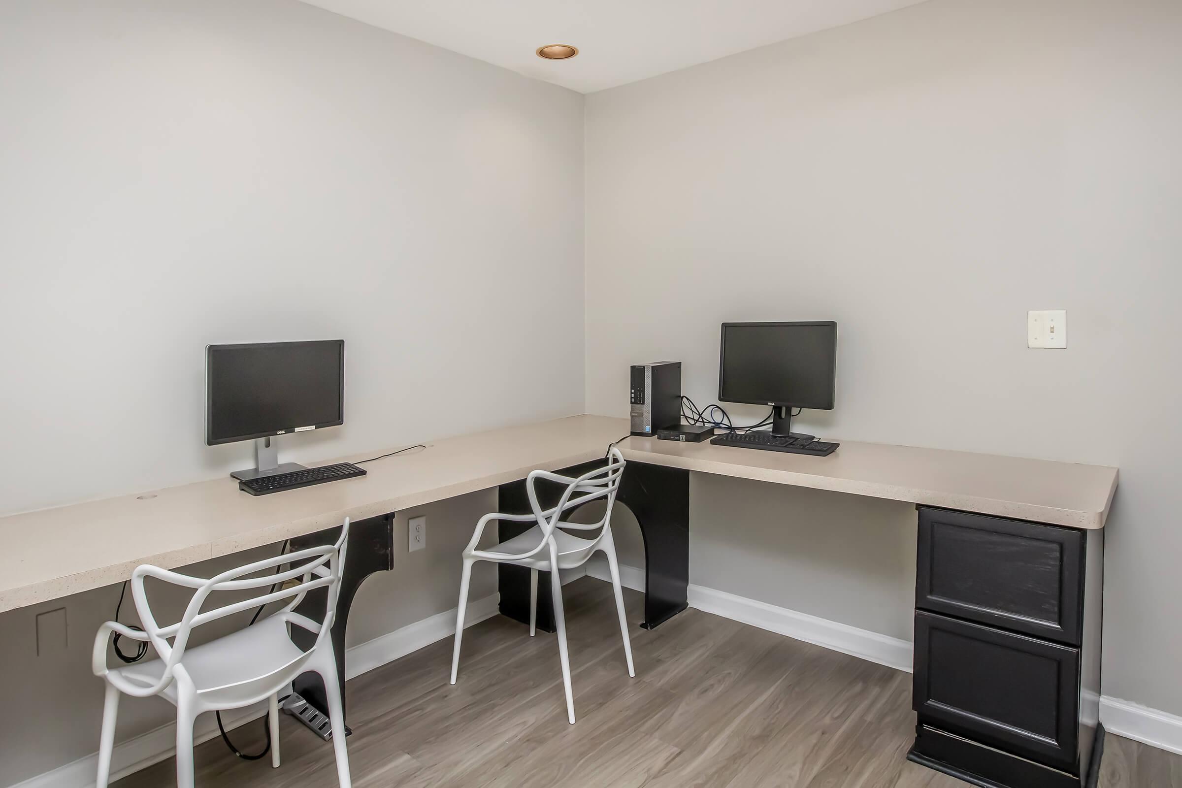 A minimalist office space featuring two desktop computers on a long, light-colored desk. There are two white, modern chairs. The walls are painted a soft gray, and the flooring is light wood. One of the computers is connected to a black CPU. The area is well-lit and organized.