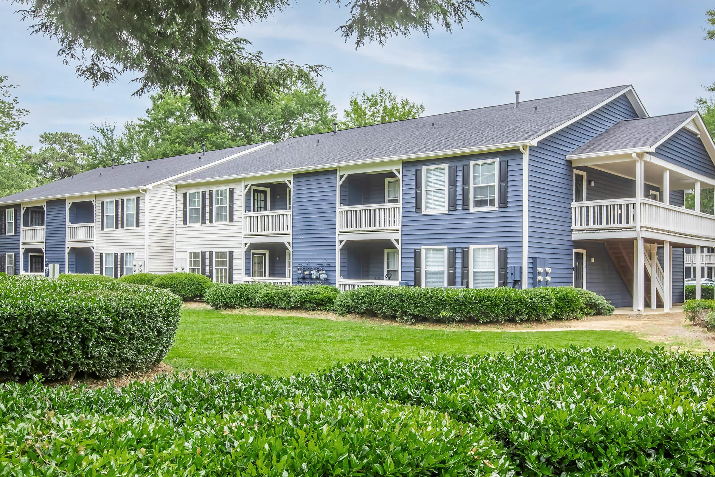 A photo of a two-story apartment complex featuring blue and white exterior walls. The building is surrounded by lush green bushes and grass, with a clear sky in the background, creating a serene residential atmosphere. Balconies are visible on each unit, adding to the inviting appearance.