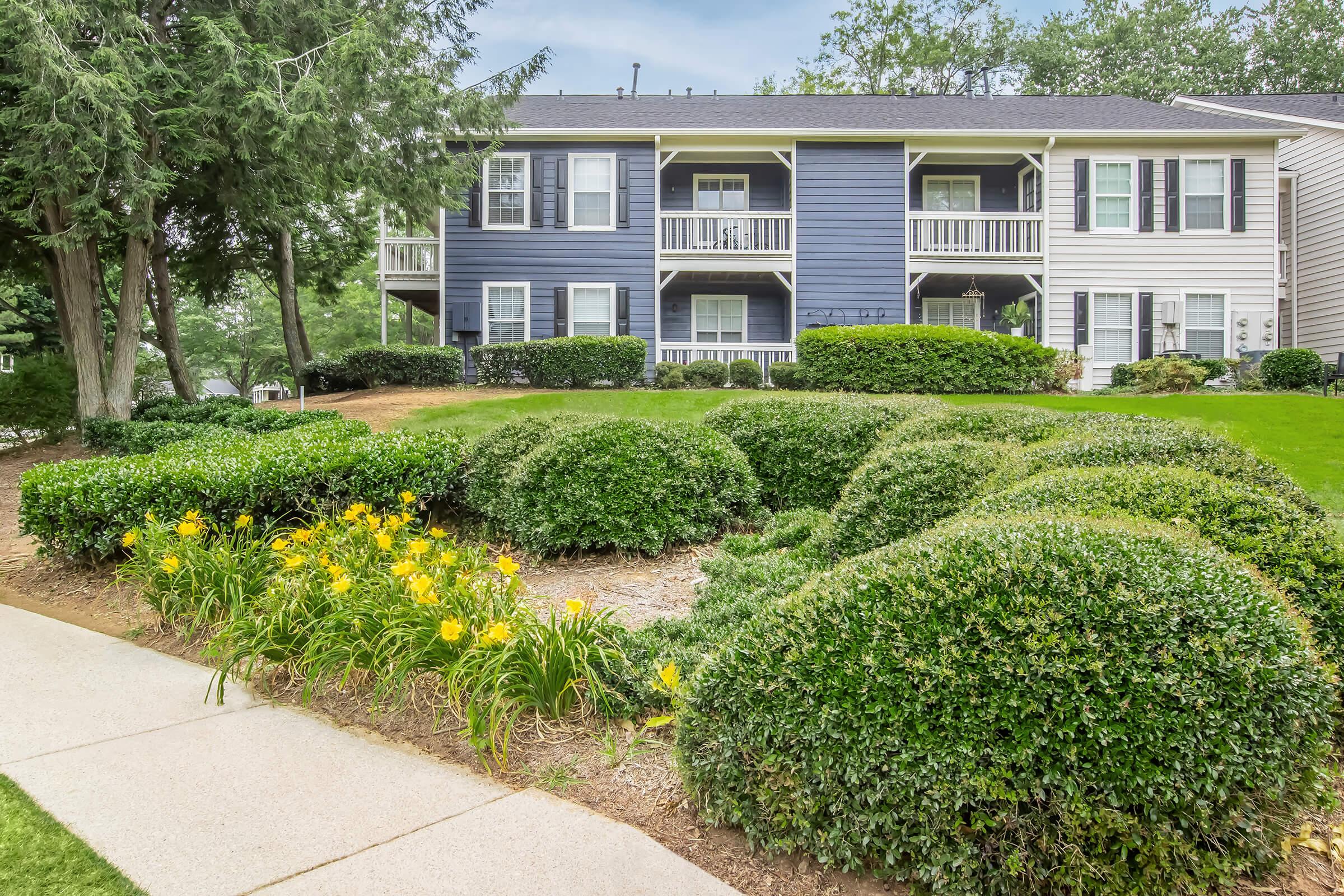 A landscaped apartment complex featuring two buildings with balconies. The foreground has manicured shrubs and vibrant yellow flowers, while the background shows a lush, green lawn and trees. The architecture includes a mix of blue and white siding.
