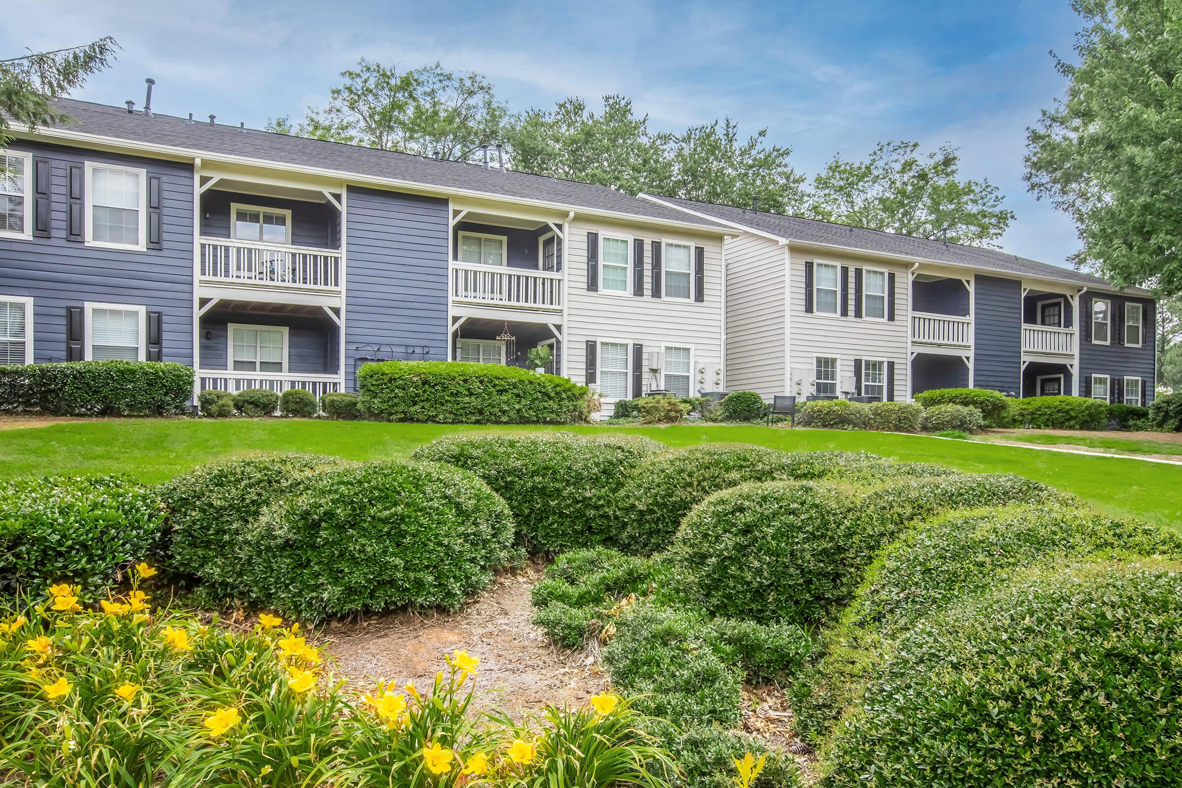 A landscaped view of a residential apartment complex with two-story buildings in shades of blue and white. The foreground features well-maintained green bushes and yellow flowers, while the background includes trees and a clear blue sky, creating a peaceful suburban atmosphere.