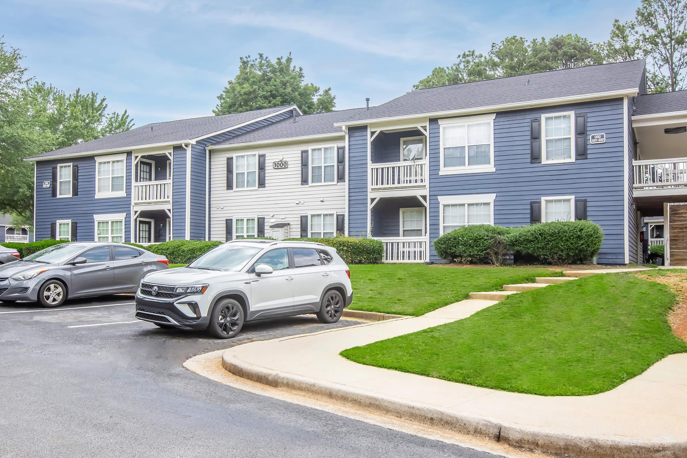 A residential apartment complex with a combination of blue and white siding. There are two buildings visible, each with balconies and greenery around. Several parked cars are in the foreground, and a pathway leads up to the entrance. The sky is cloudy, suggesting a calm atmosphere.