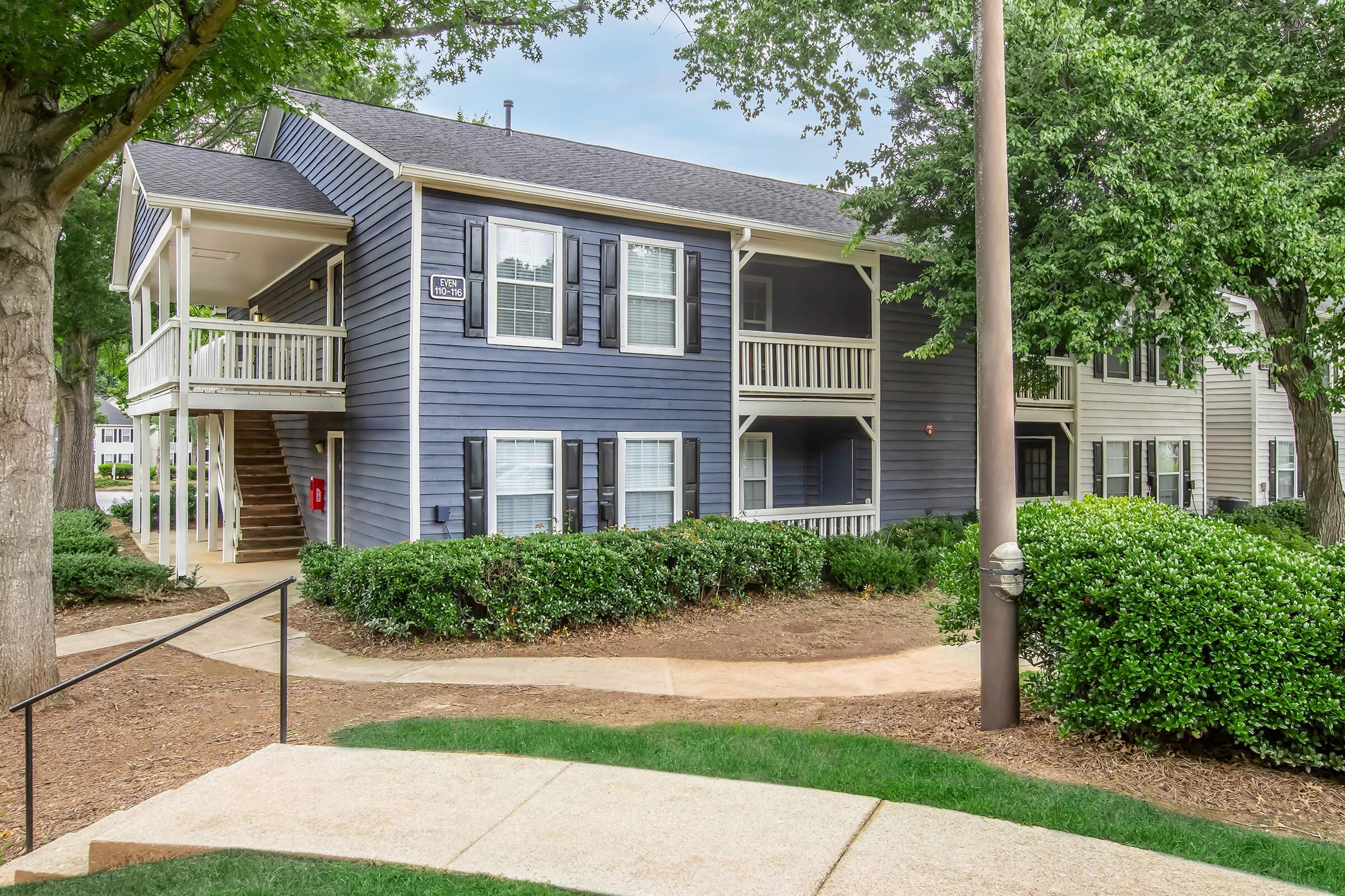 Two-story residential building with blue siding and white trim. Features multiple windows, a front porch, and a landscaped path leading up to it. Surrounded by green shrubs and trees, the building is set in a well-maintained area with a sidewalk and other similar buildings nearby.