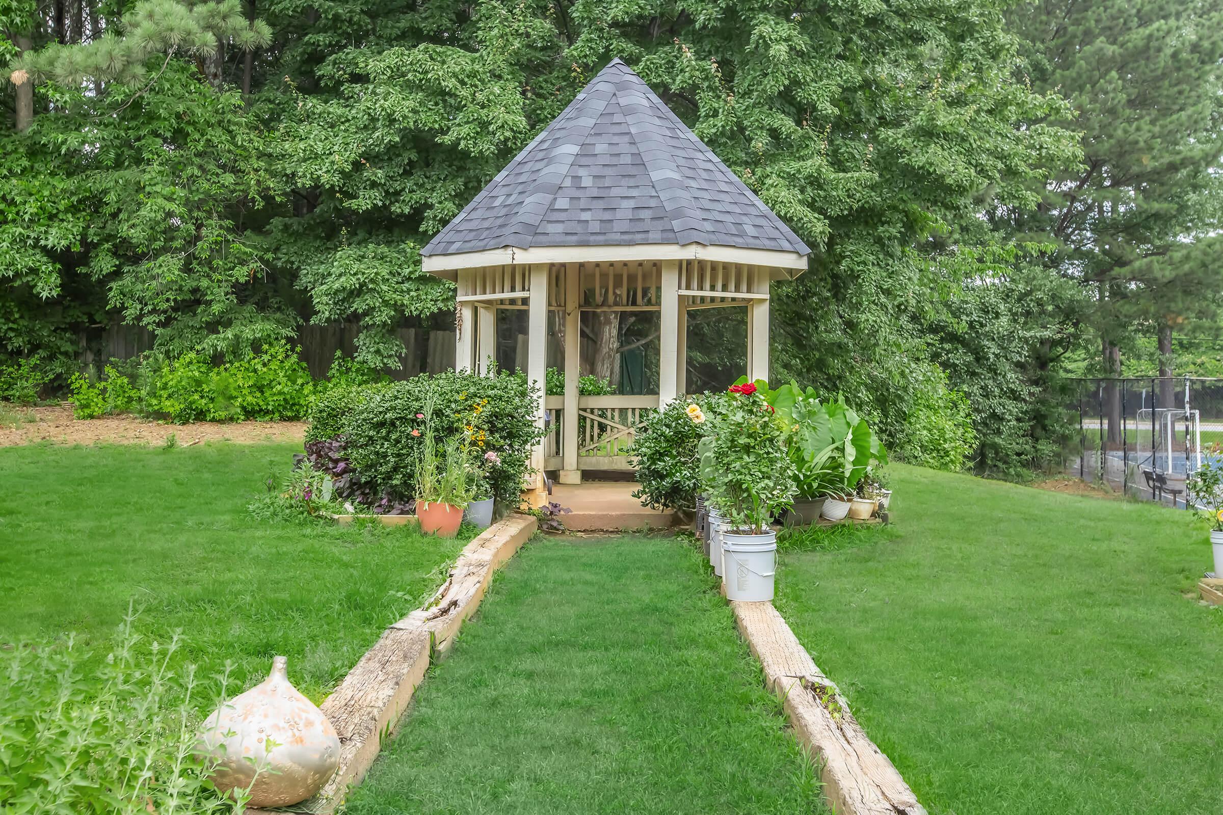 A quaint, white gazebo with a pointed roof sits at the end of a green lawn, surrounded by lush trees and various plants. A pathway lined with potted flowers leads to the gazebo, creating a picturesque garden setting. The area is peaceful and well-maintained, ideal for relaxation or gatherings.