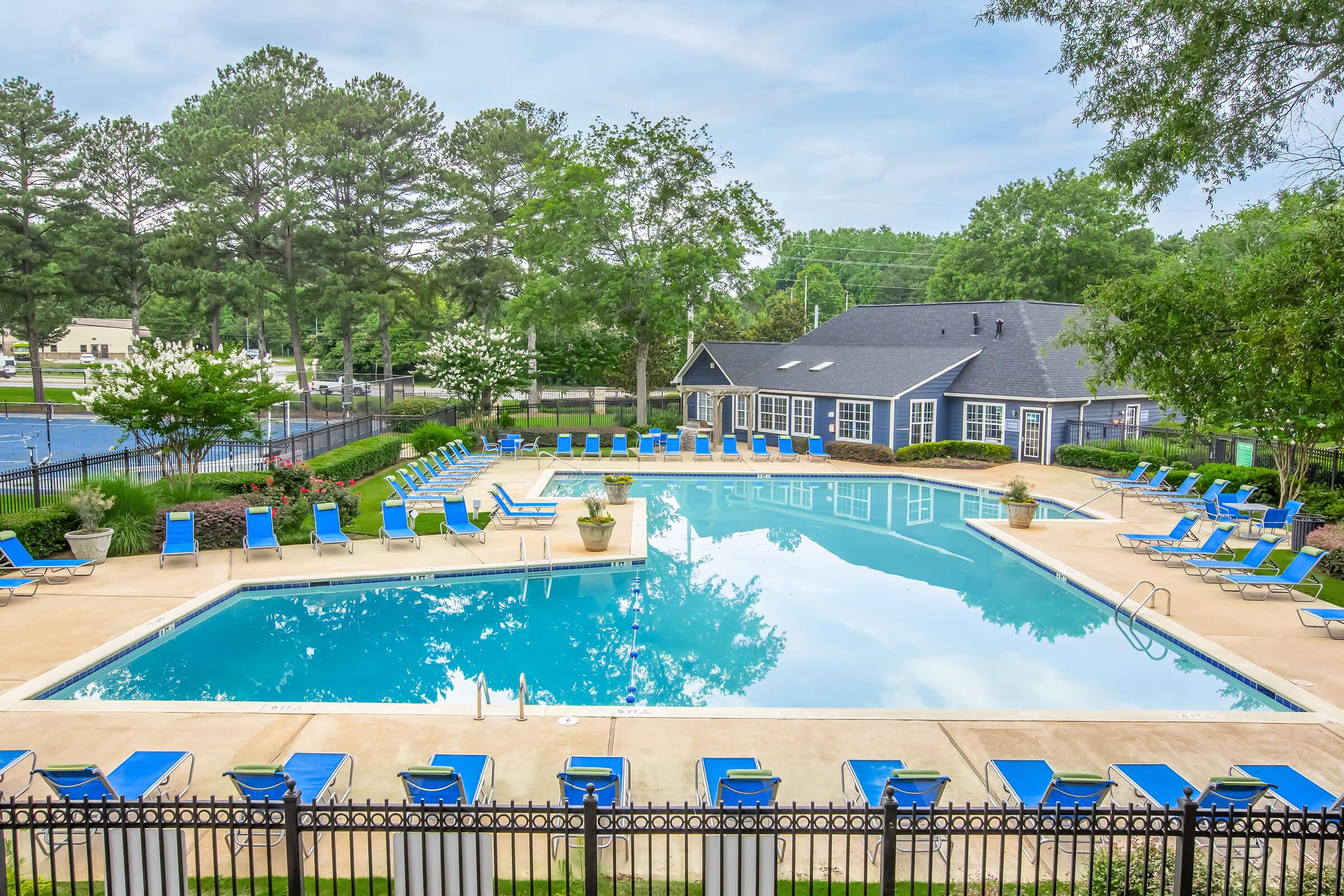 A serene pool area featuring a large, clear swimming pool surrounded by lounge chairs. Lush greenery and potted plants enhance the scenic view, with a clubhouse nearby. Tennis courts are visible in the background, creating a relaxing outdoor space.