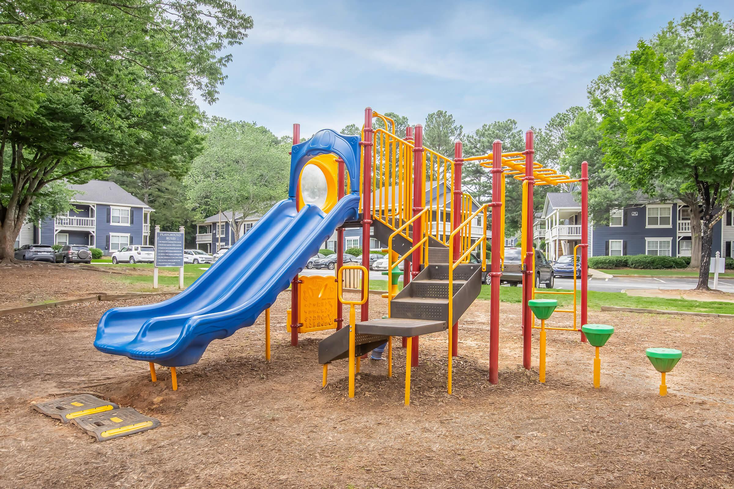 A colorful playground featuring a blue slide, climbing structure, and various play features. The area has a soft ground surface and is surrounded by trees and residential buildings in the background. The scene is inviting and designed for children’s play and exploration.