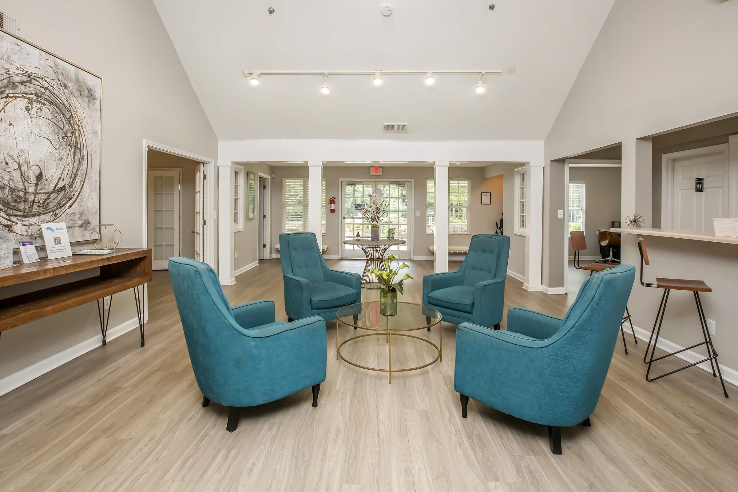 A modern lobby area featuring four teal armchairs arranged around a circular glass coffee table. The space has wooden flooring and light-colored walls, with a large artwork on one side and a reception area visible in the background, creating an inviting atmosphere. Natural light comes from large windows.