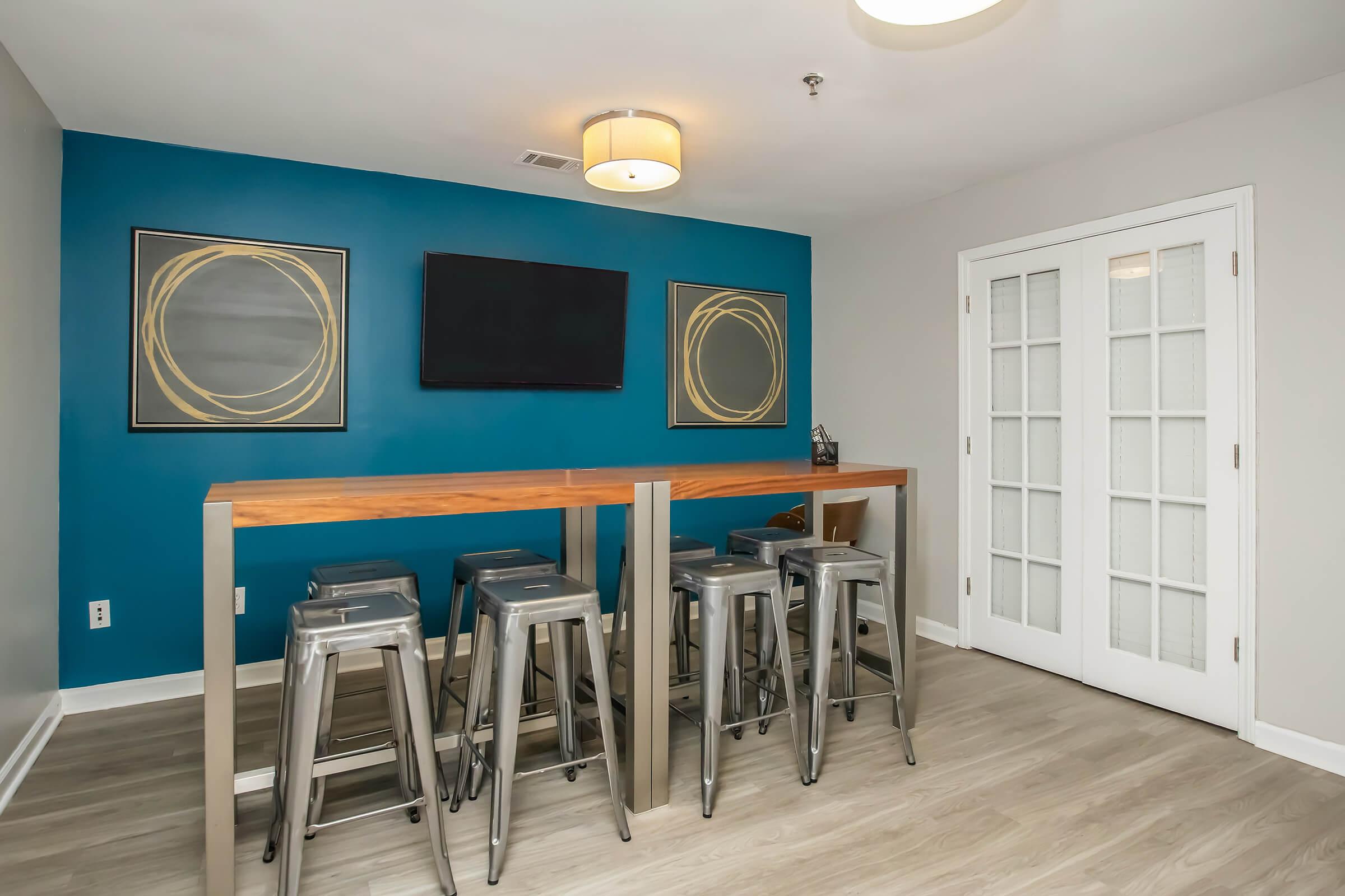 A modern room featuring a blue accent wall with two abstract circular artworks, a flat-screen TV mounted above a sleek wooden bar table with metal stools. Glass double doors on the right lead to another space, and the floor is finished with light-colored wood.