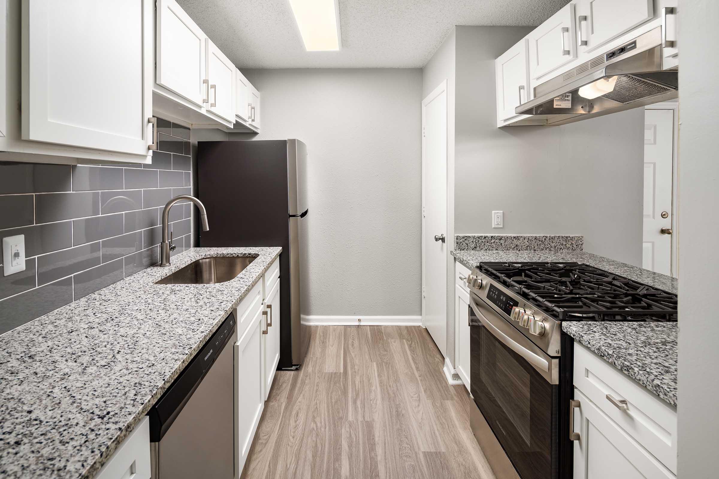 Modern kitchen featuring white cabinetry, granite countertops, stainless steel appliances including a refrigerator and gas stove, and a sleek backsplash. The room has ample natural light and a neutral color palette, with a spacious layout designed for functionality.