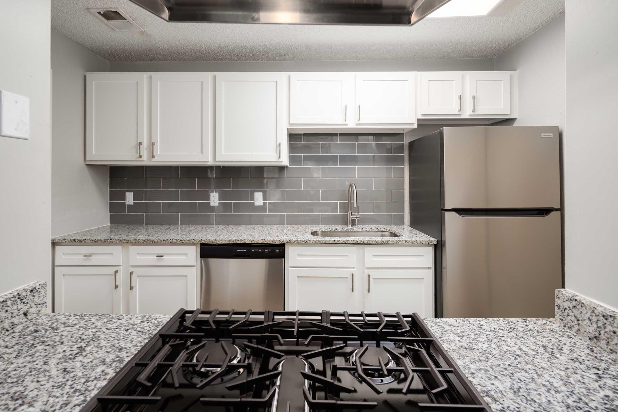Modern kitchen featuring white cabinets, stainless steel appliances, and a granite countertop. The space includes a refrigerator, dishwasher, and a sleek gas stove. The backsplash is made of gray tiles, adding a contemporary touch to the overall design.