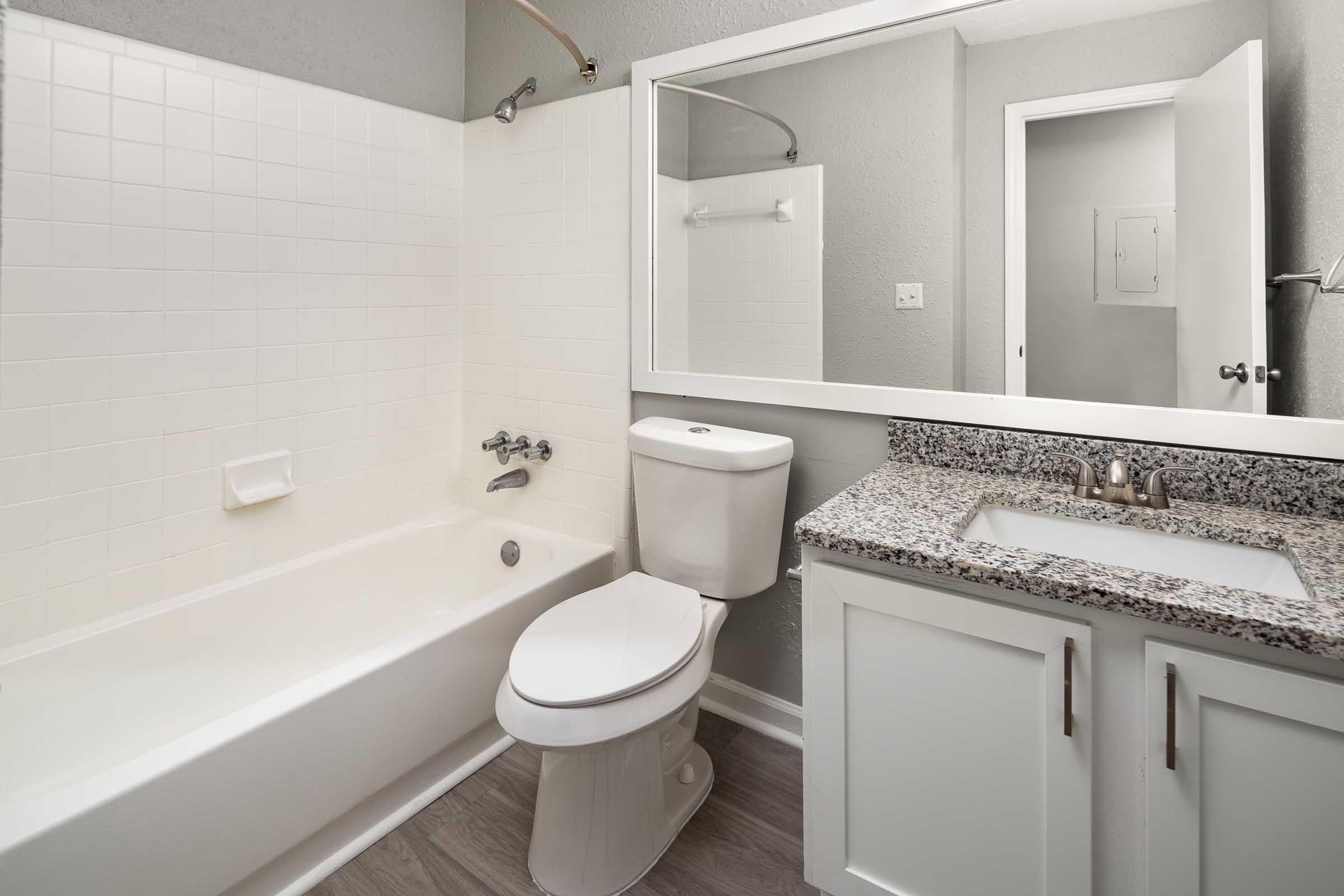 A modern bathroom featuring a white bathtub, a toilet, and a granite countertop with a sink and mirror. The walls are painted light gray, and there's a doorway visible in the background. The overall design is clean and contemporary.