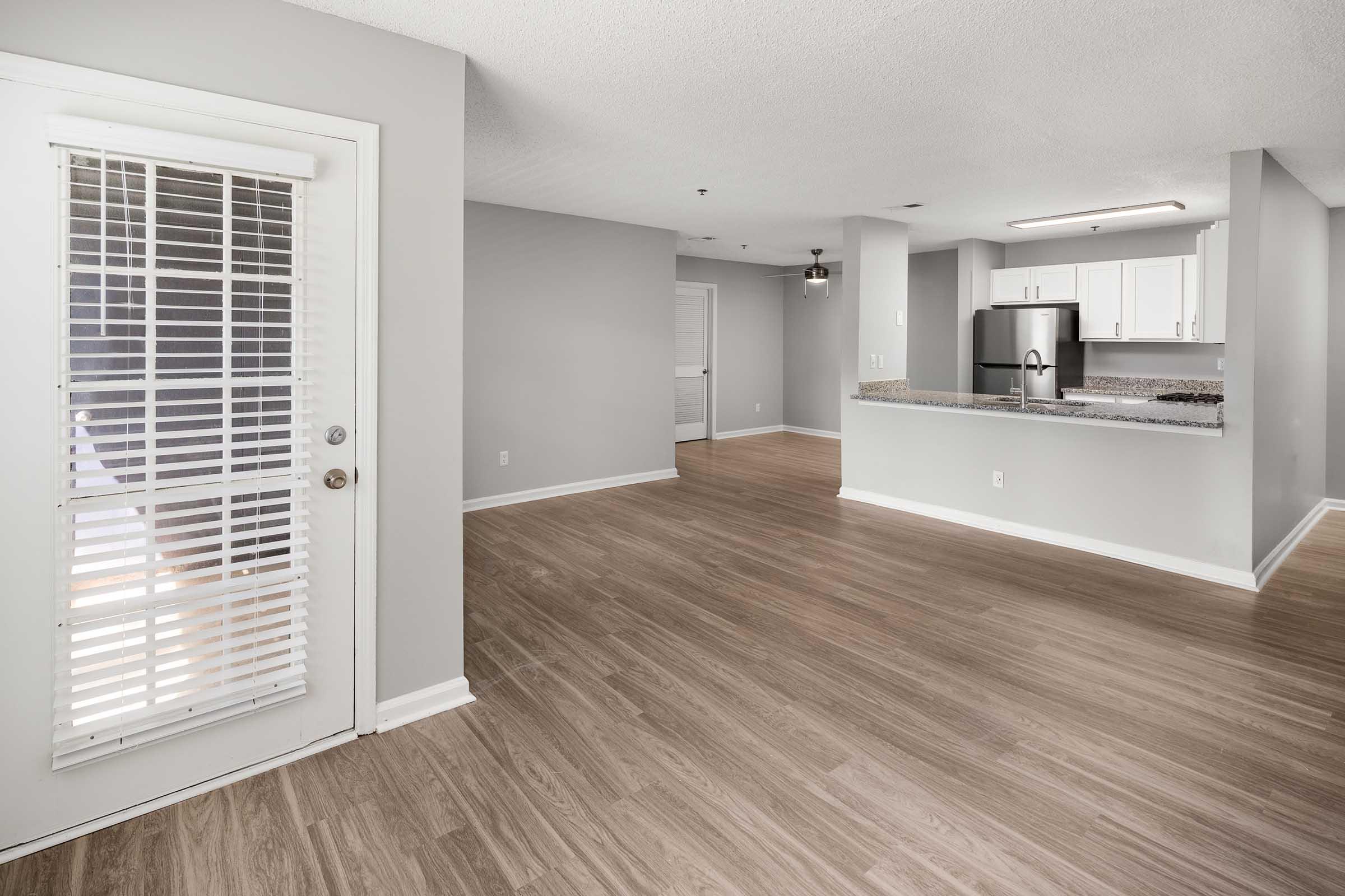 Bright, open living space with light gray walls and wood-style flooring. A door with white blinds leads outside, and the kitchen features modern white cabinetry and a granite countertop. The area is well-lit, creating a welcoming atmosphere.