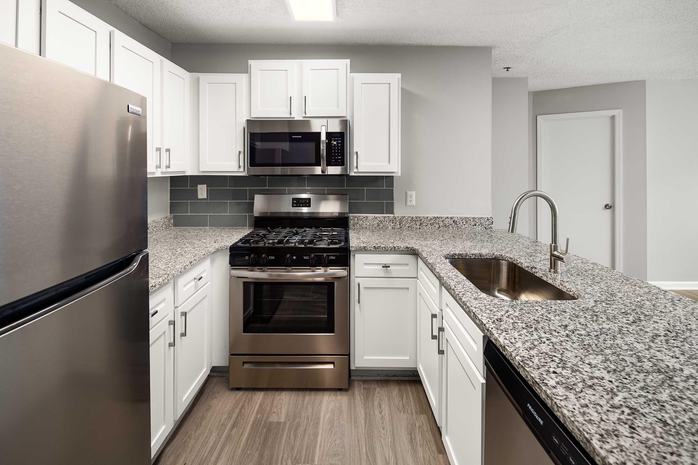Modern kitchen featuring stainless steel appliances, including a refrigerator, stove, and microwave. White cabinets complement a gray backsplash and granite countertops. A single basin sink is integrated into the countertop, with wooden flooring visible in the foreground.