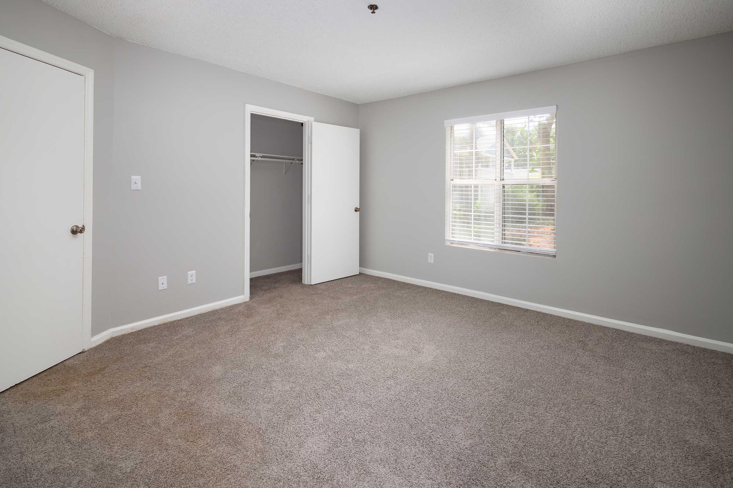 A spacious, empty bedroom featuring light gray walls, carpeted flooring, and a window allowing natural light. The room includes a closet with a white door, another door leading to an adjoining space, and no furniture or decorations present.