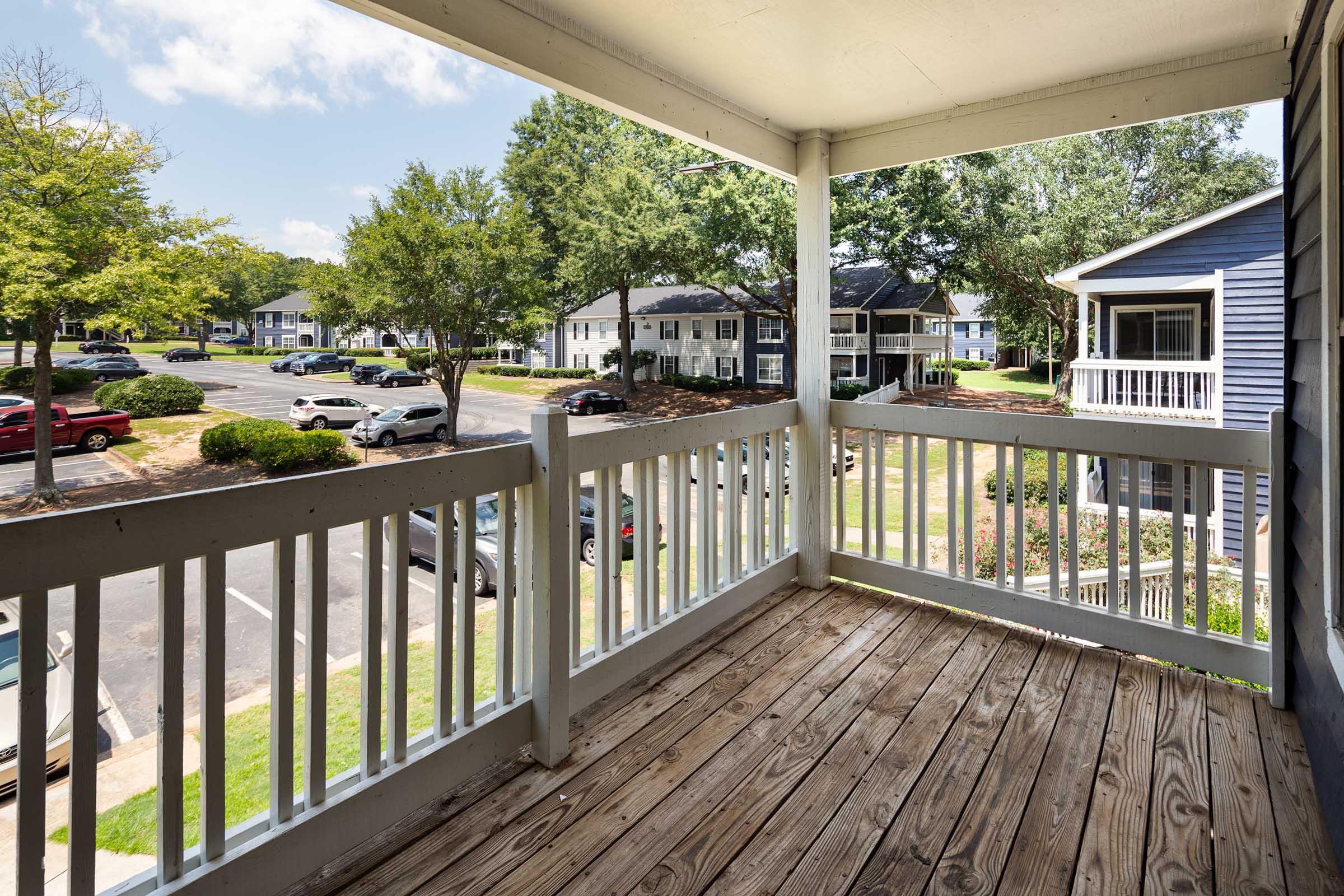View from a balcony overlooking a neighborhood with trees and parked cars. The scene features a mix of buildings, including two-story apartments and well-maintained lawns, under a partly cloudy sky.