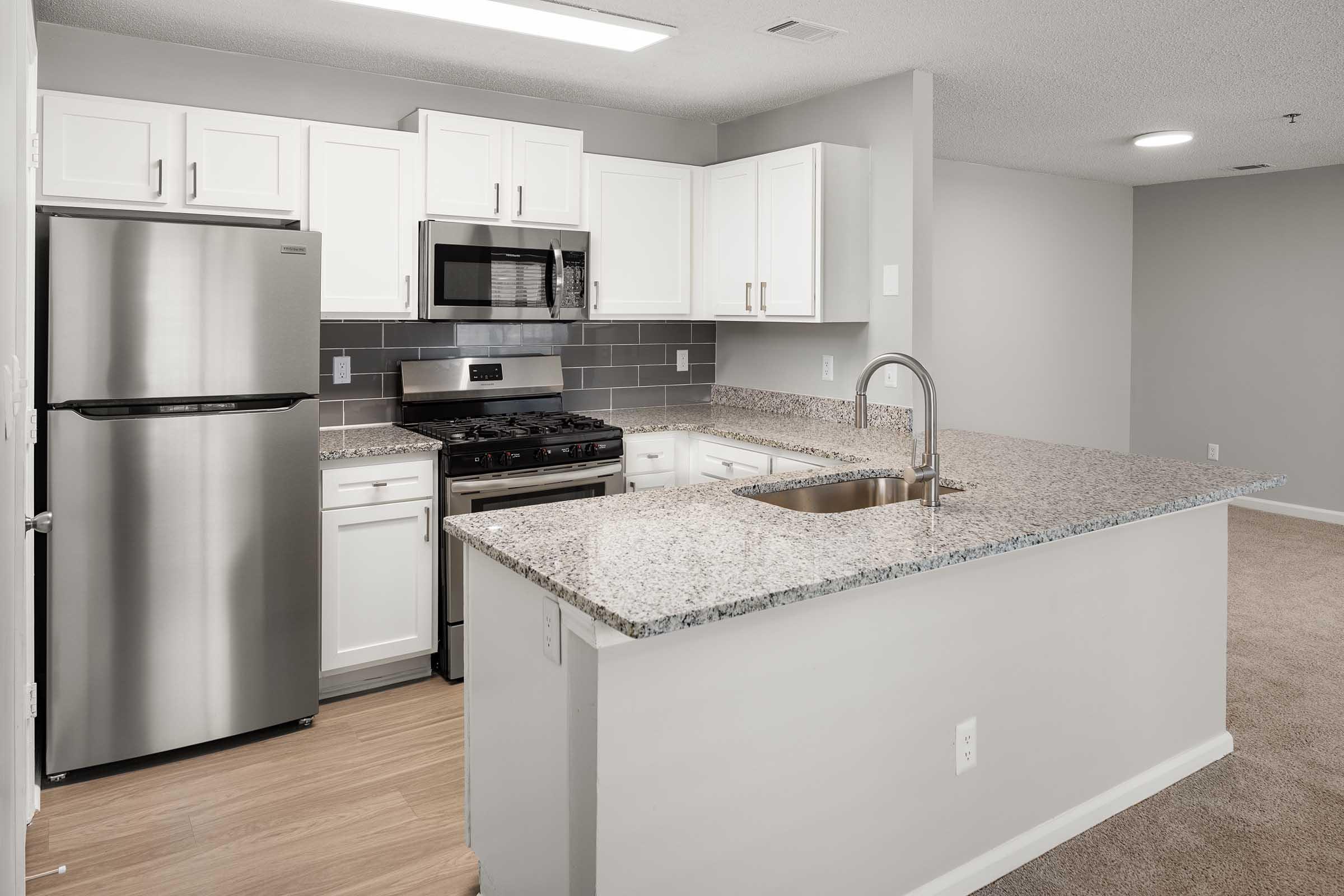 Modern kitchen featuring gray granite countertops, white cabinetry, stainless steel appliances including a refrigerator and microwave, and a gas stove. The open layout connects the kitchen to a living area with beige carpeting. Brightly lit with natural light from a window above the sink.