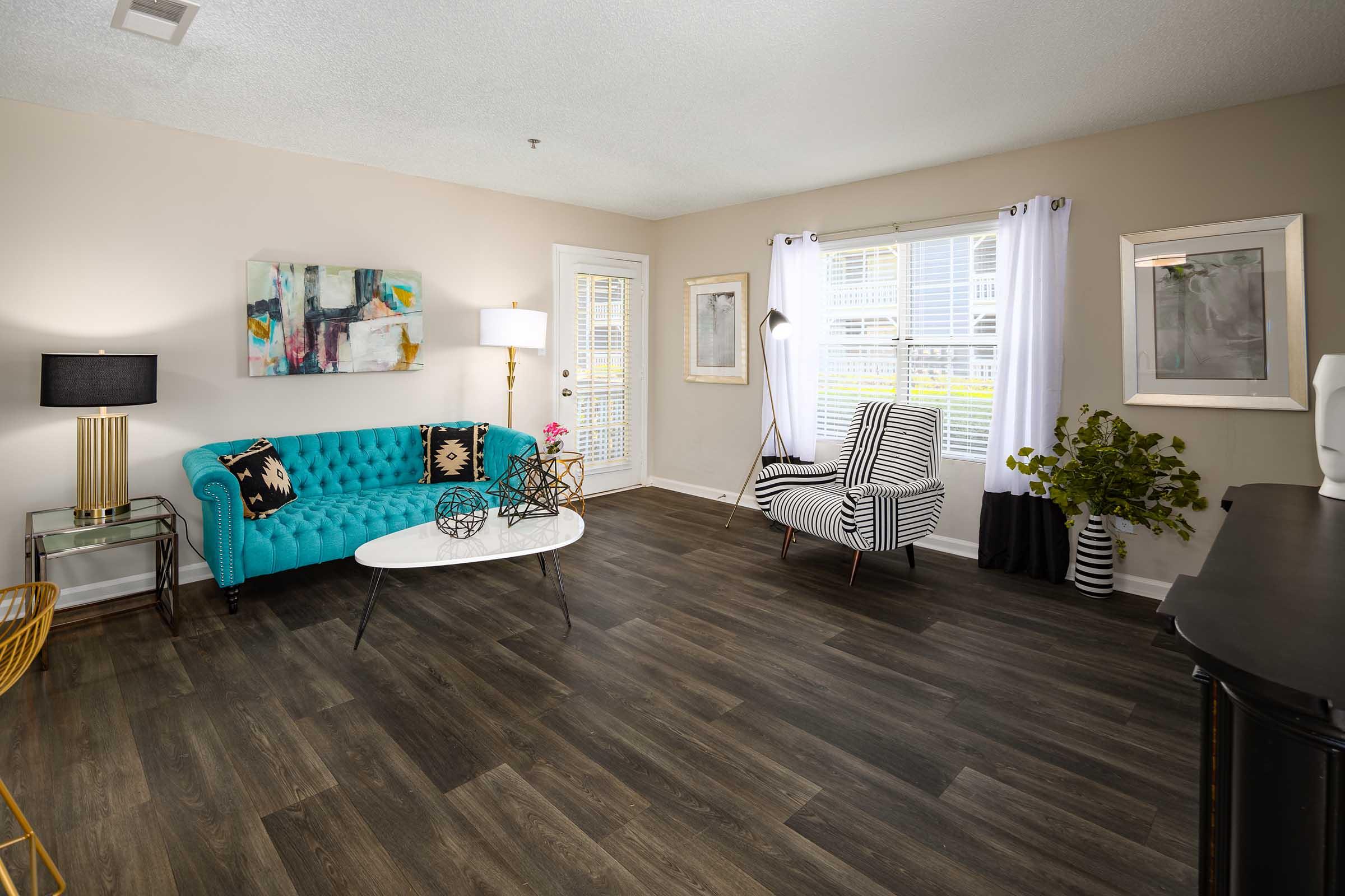 A modern living room featuring a turquoise tufted sofa, a white coffee table, and a striped accent chair. The space includes a lamp, decorative plants, and artwork on the walls, with large windows allowing natural light to illuminate the room. The flooring is dark wood, enhancing the contemporary aesthetic.