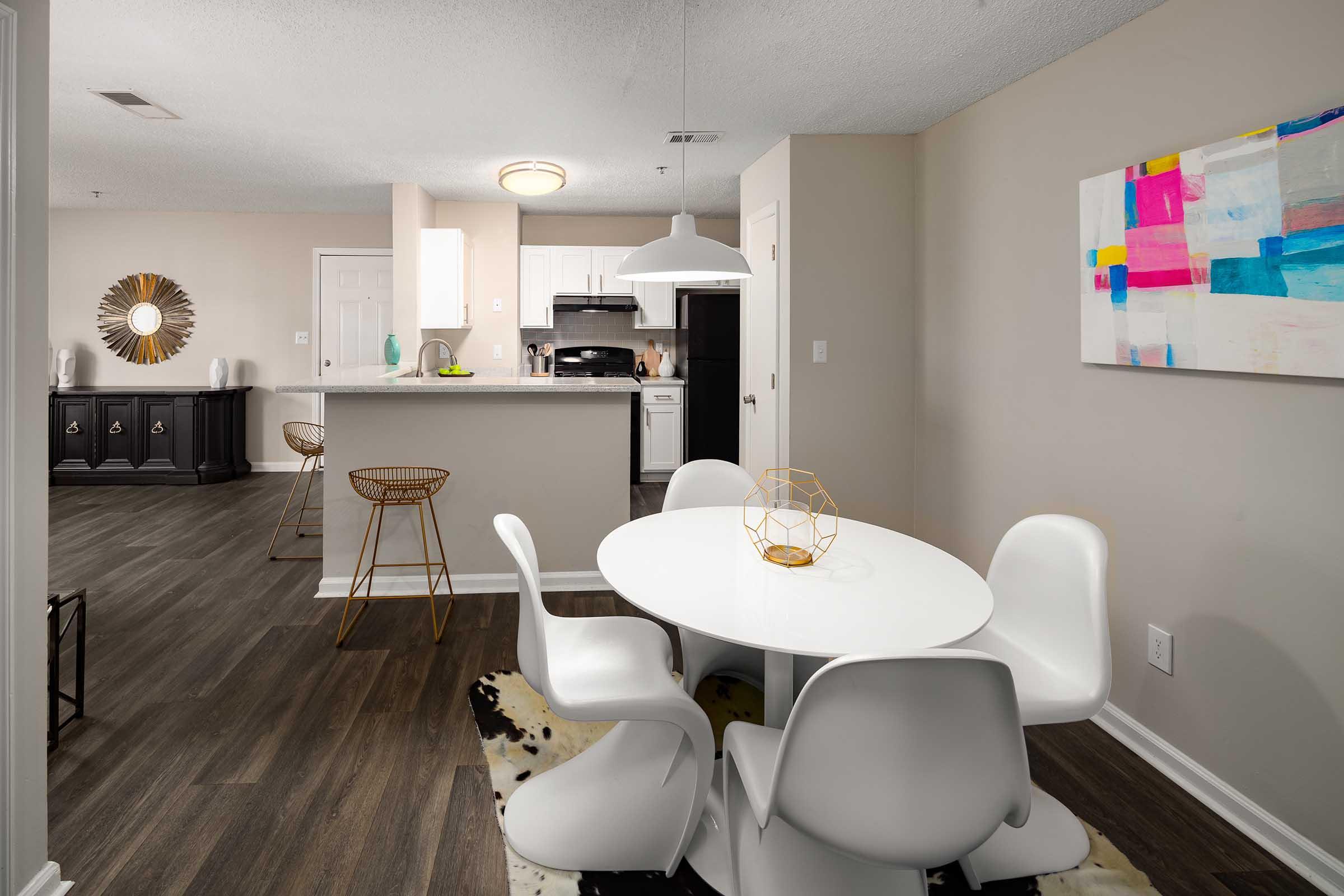 A modern dining area featuring a round white table with four white chairs, a geometric decor piece on the table, and a stylish rug. The open kitchen can be seen in the background, equipped with stainless steel appliances. A colorful abstract painting hangs on the wall, adding vibrancy to the neutral-toned interior.