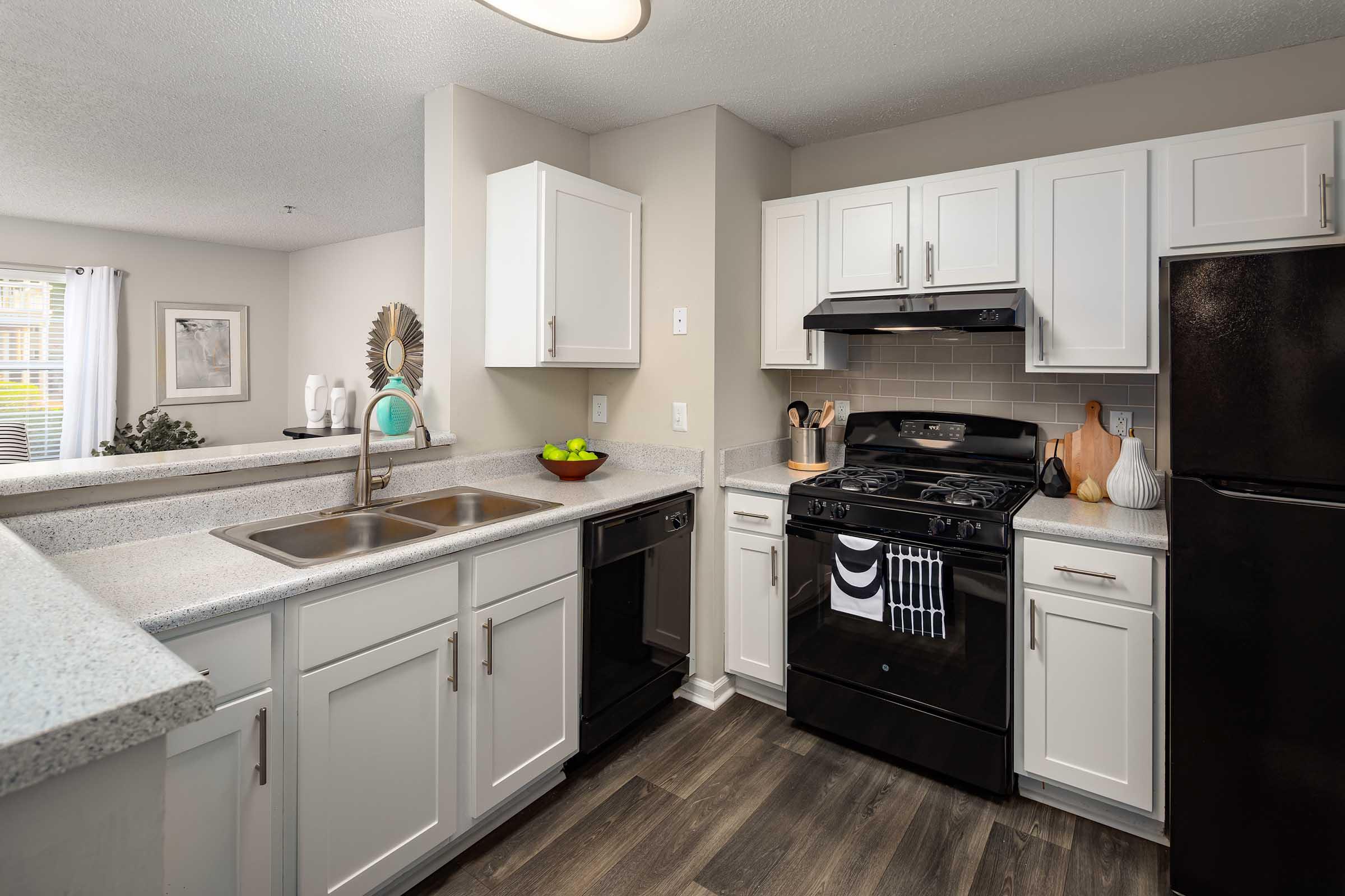Modern kitchen featuring white cabinetry, a double sink, a black gas stove, and an island with gray countertops. Appliances include a black refrigerator and a dishwasher. The space is well-lit with natural light coming from a nearby window, complemented by decorative elements and a bowl of fruit on the countertop.